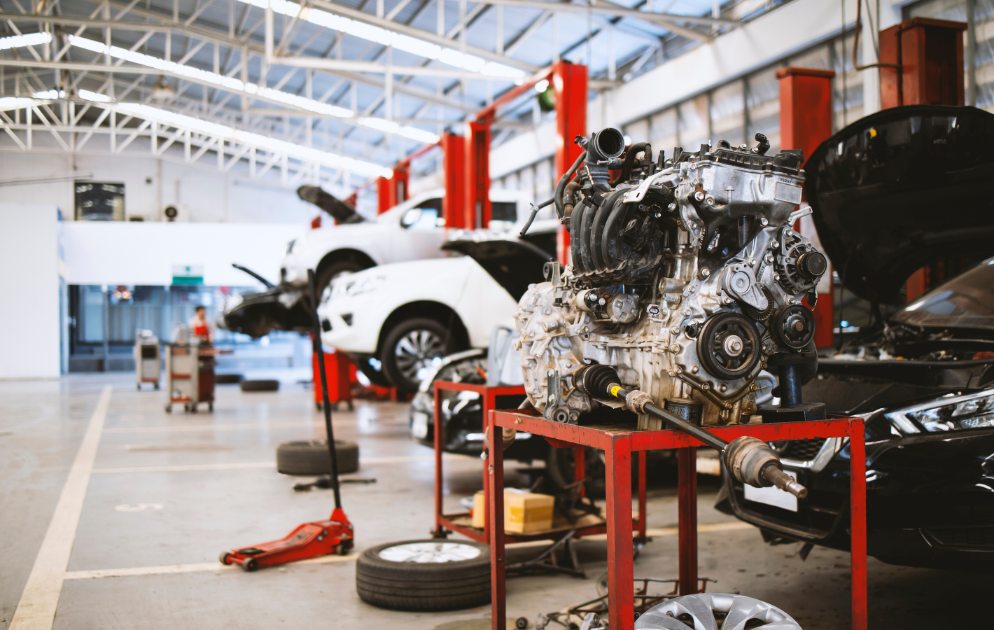Car engine on a red stand in a bright auto repair shop with several cars on lifts.