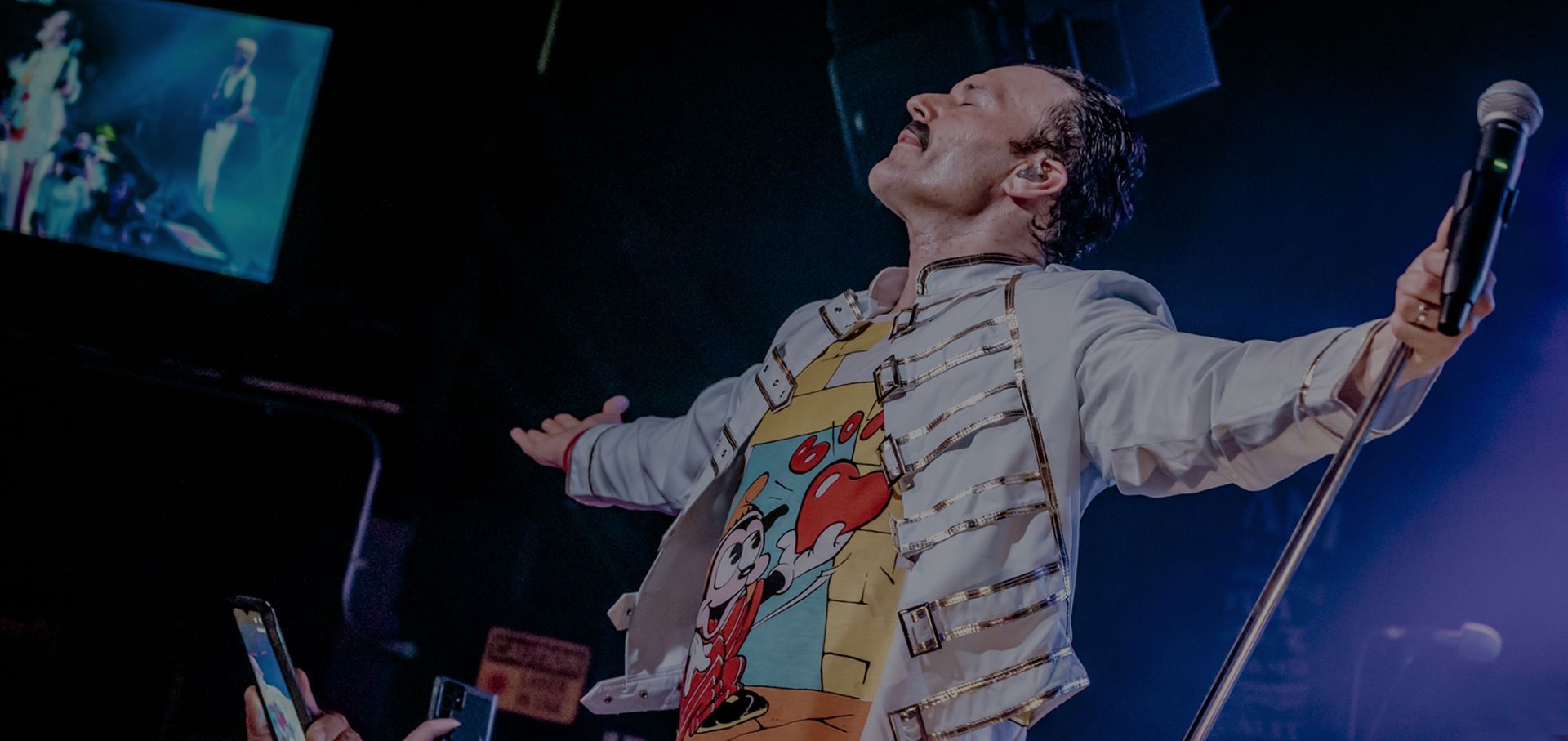 Singer on stage with arms outstretched, holding a microphone, under blue concert lighting.