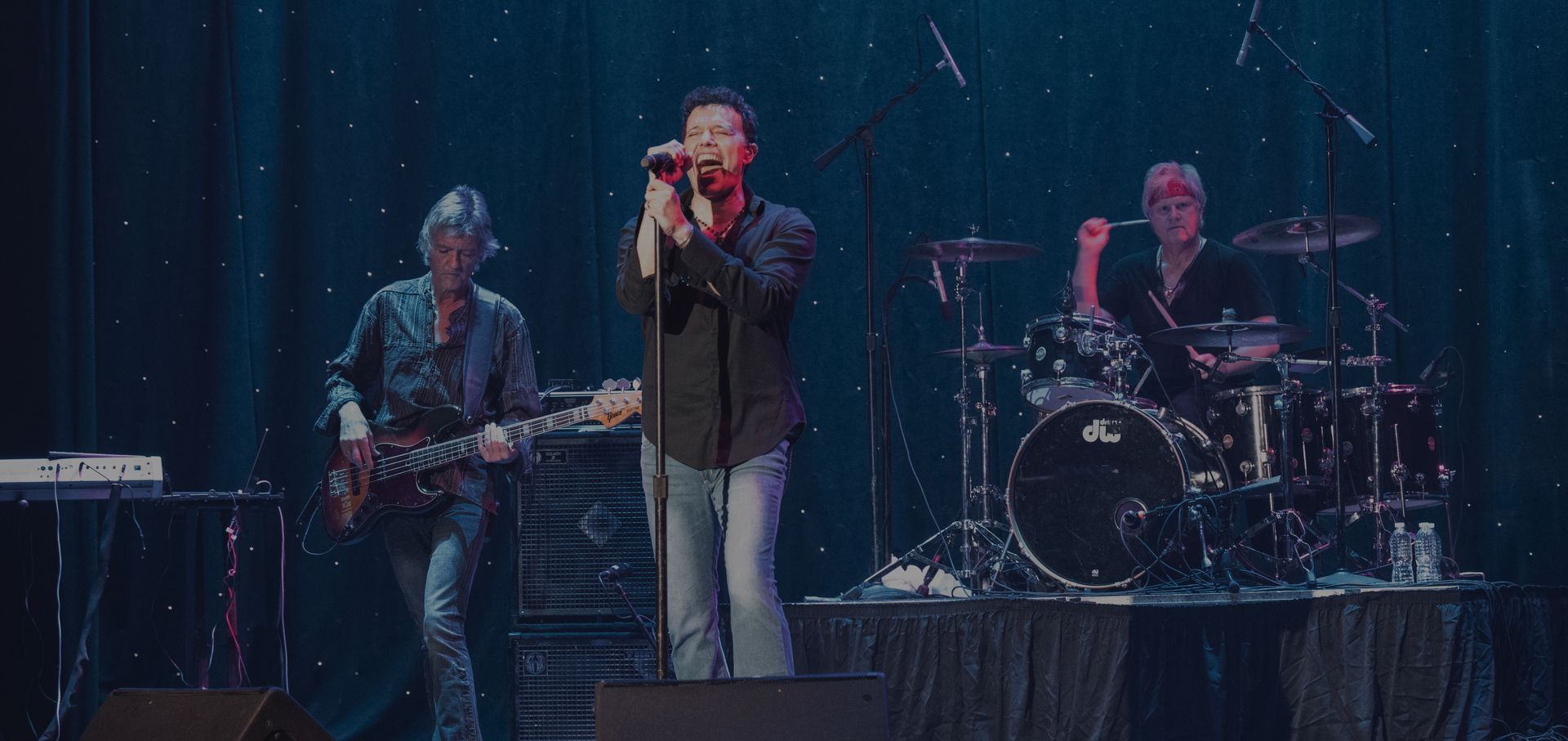 Three-piece band performing on stage under blue lights, with singer at center and drummer in back.