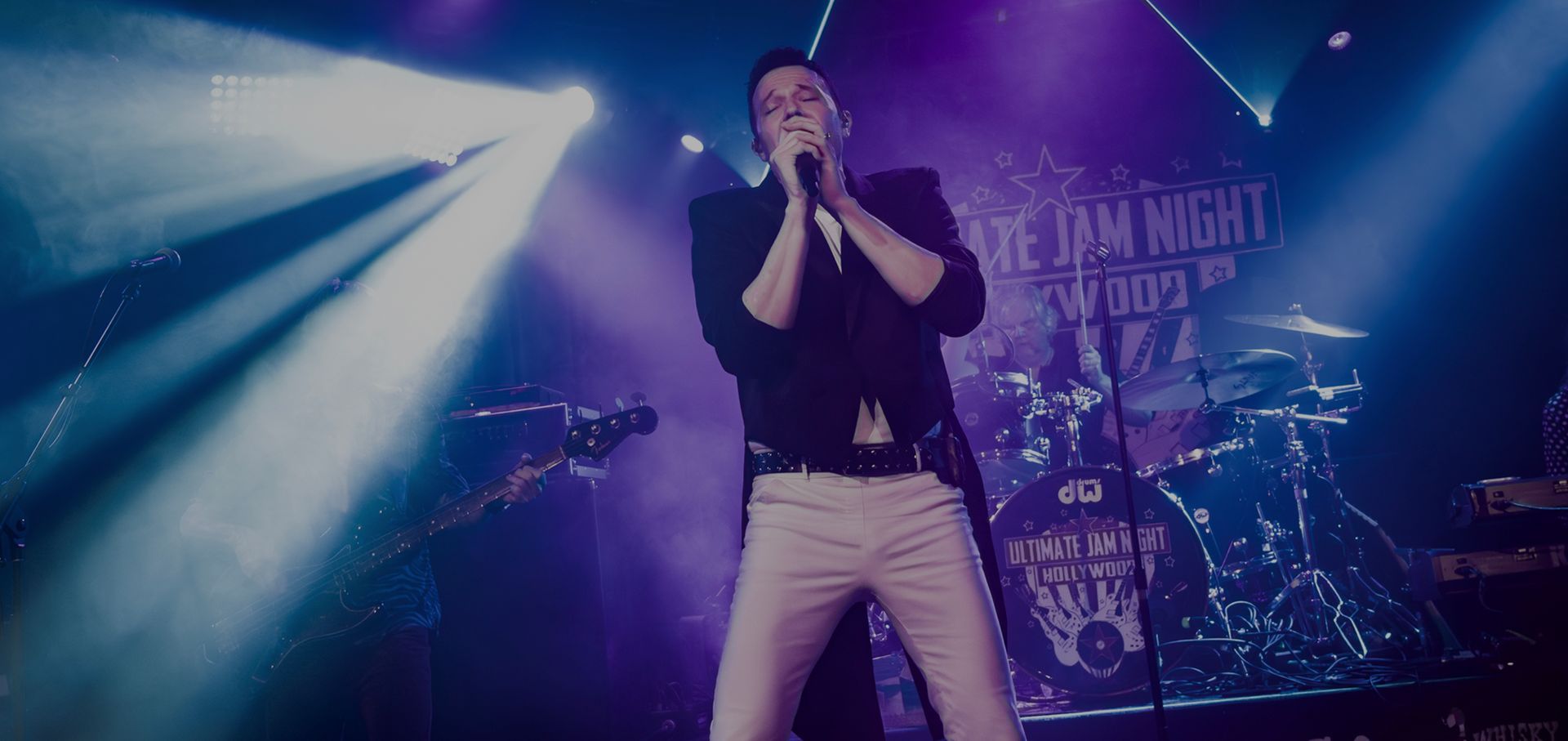 Singer performing on a purple-lit stage, holding a microphone under bright spotlights.