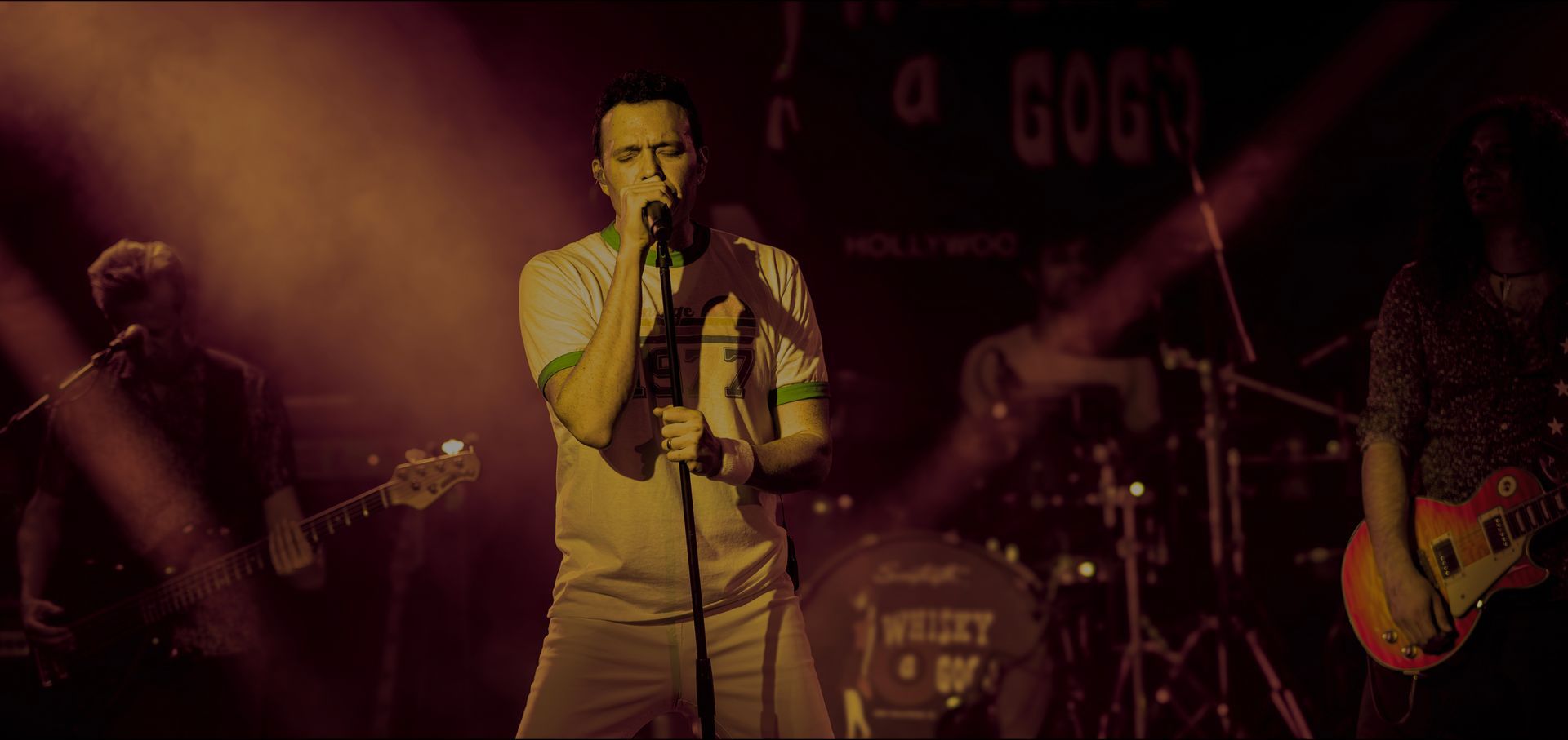 Singer on stage with microphone under warm red lighting, band instruments visible in the background