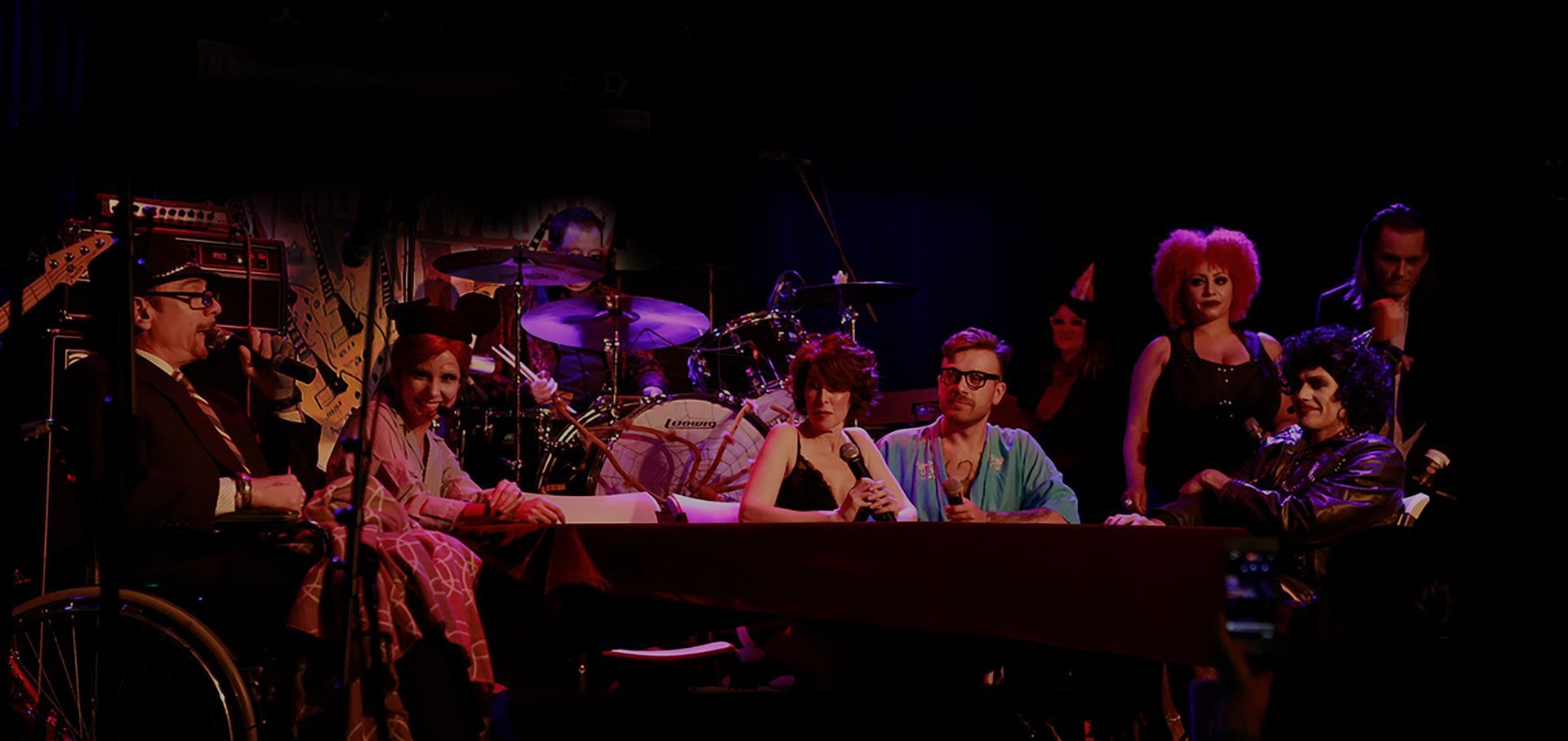 Costumed performers seated at a table on a dimly lit stage, with a bright pink wig visible on the right
