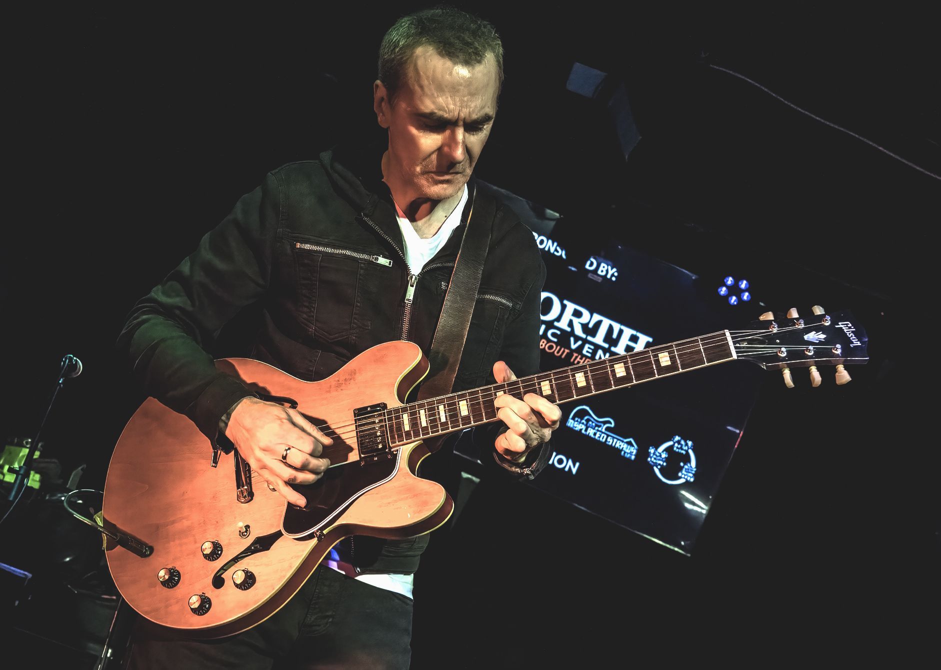 Man playing a hollow-body electric guitar onstage, lit by stage lights.