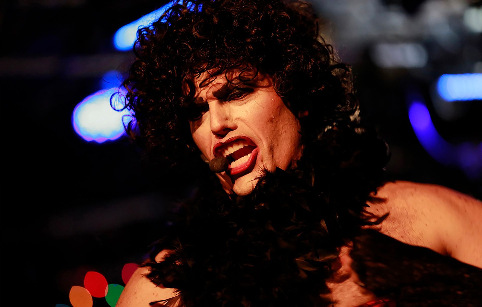 A man with curly hair is wearing a black feather boa around his neck while singing