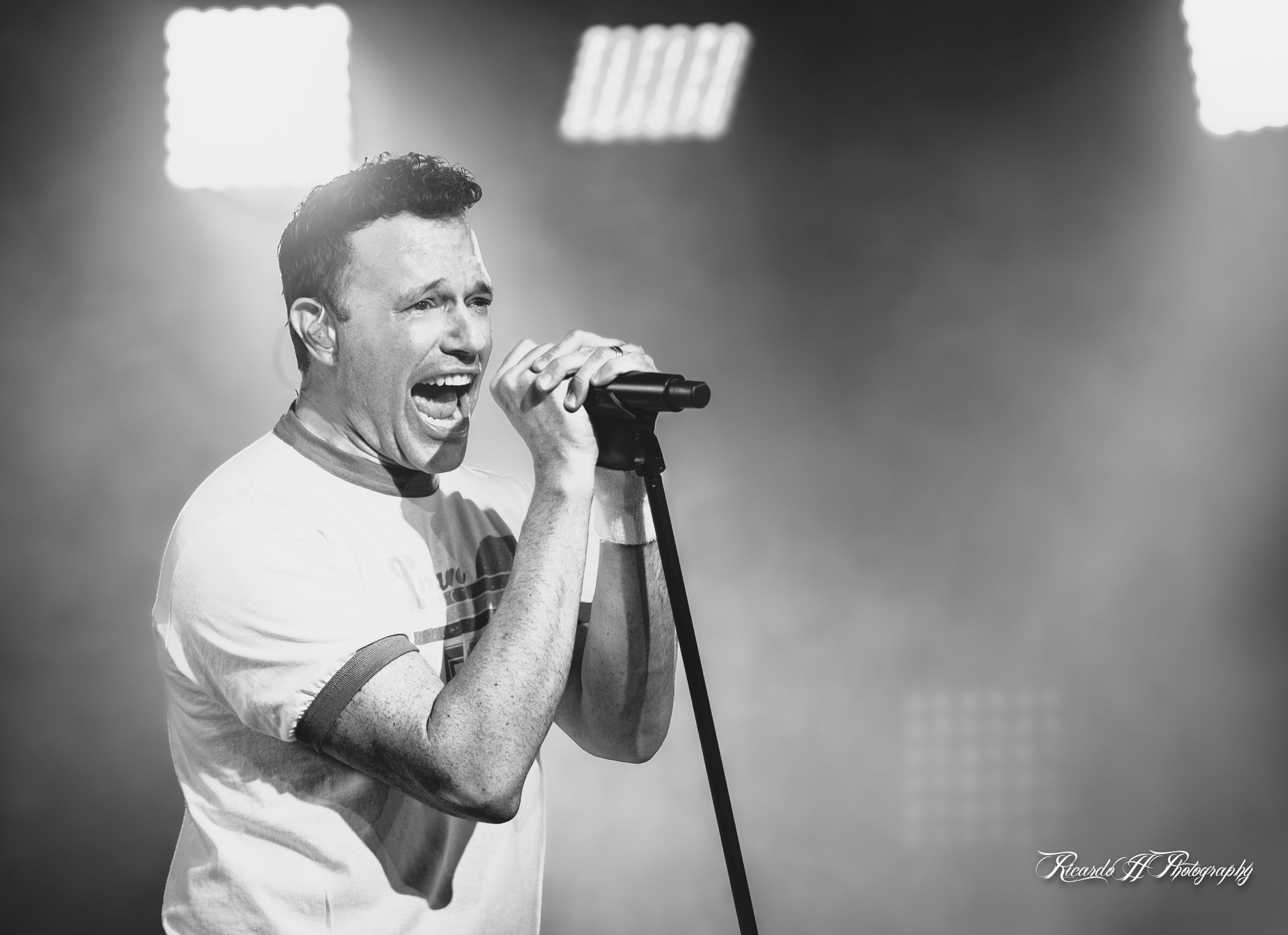 A man is singing into a microphone on a stage in a black and white photo.