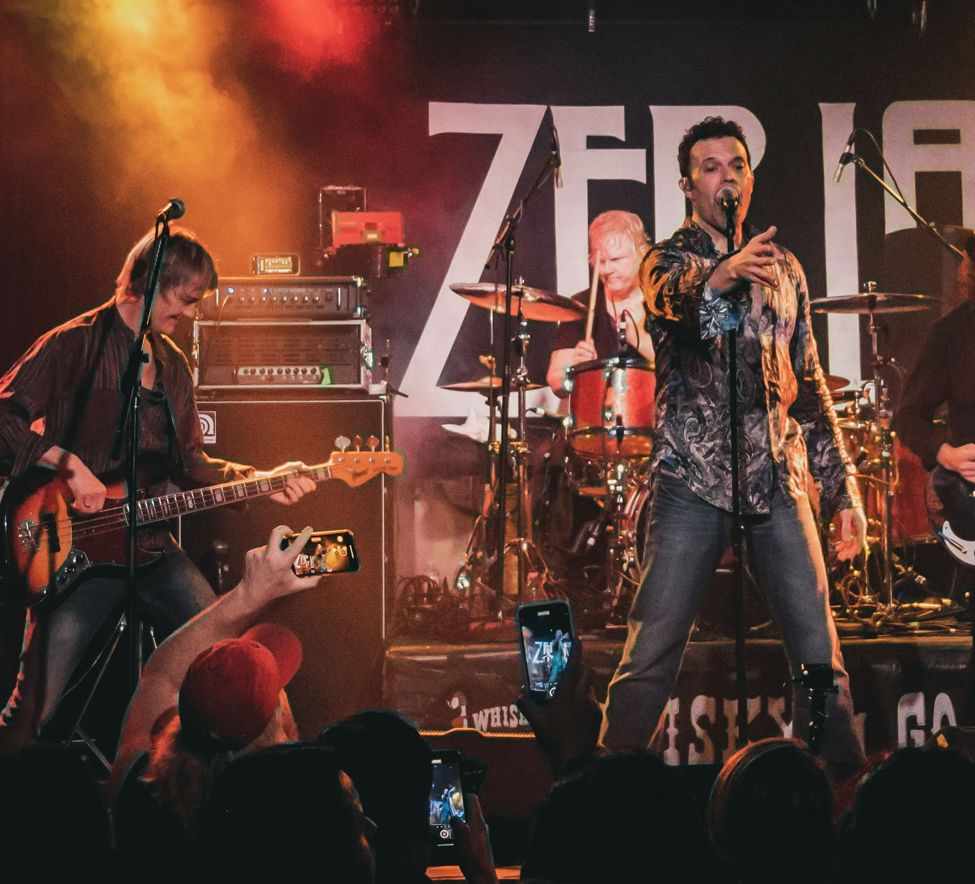 Band performing on stage, singer with microphone, drummer, bassist, audience taking photos.