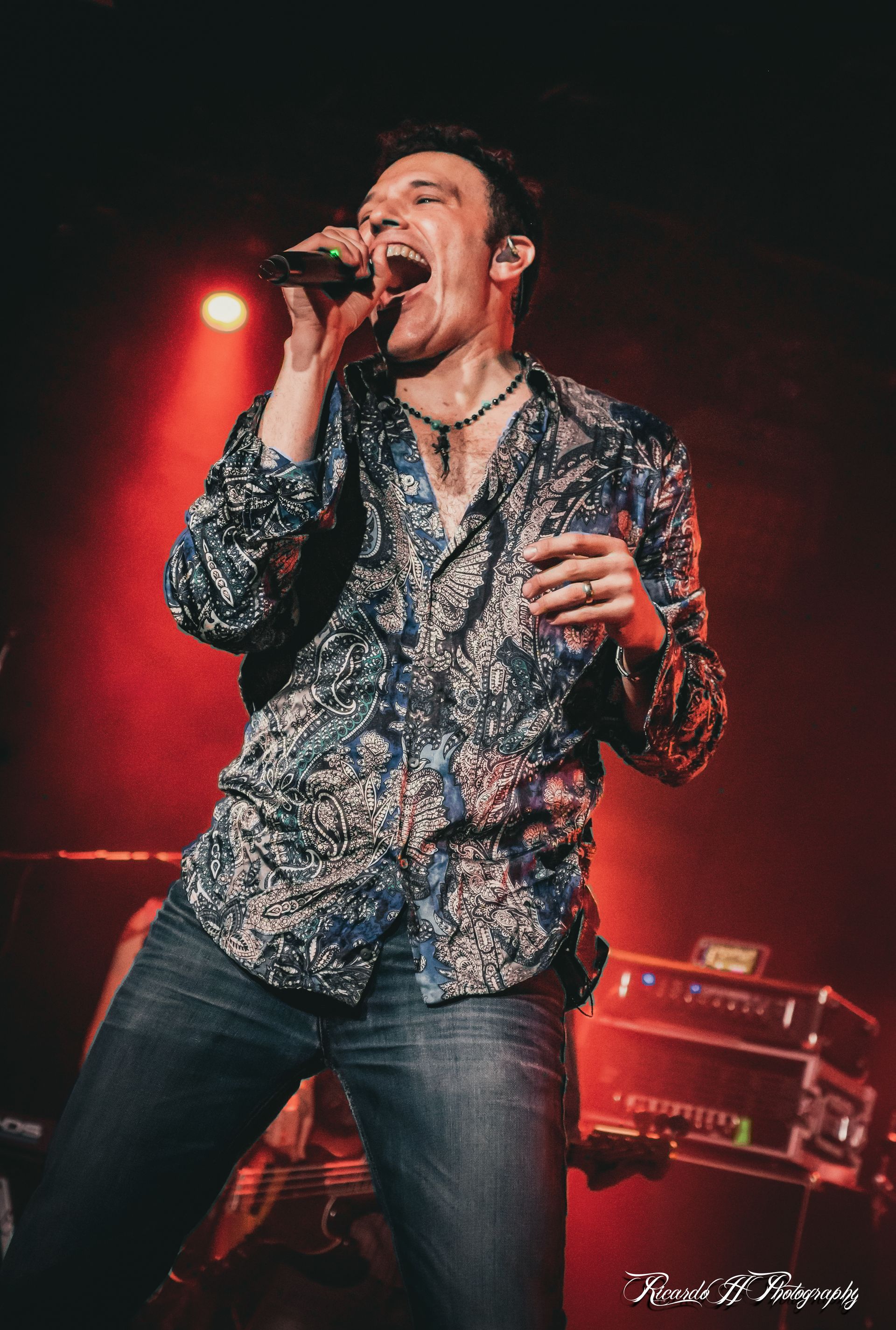 Man singing into a microphone onstage, lit by red light. He wears a blue patterned shirt and jeans, singing with an open mouth.
