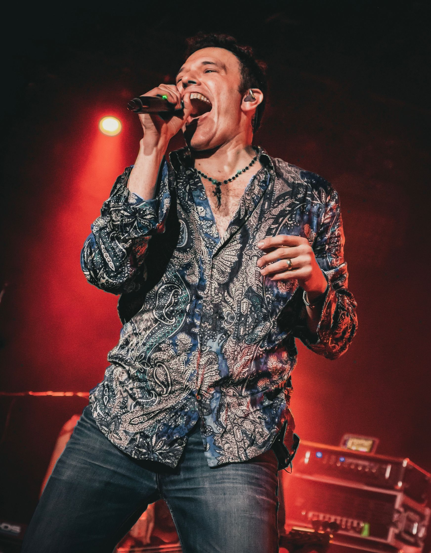 Man singing into a microphone onstage, lit by red light. He wears a patterned shirt and jeans.