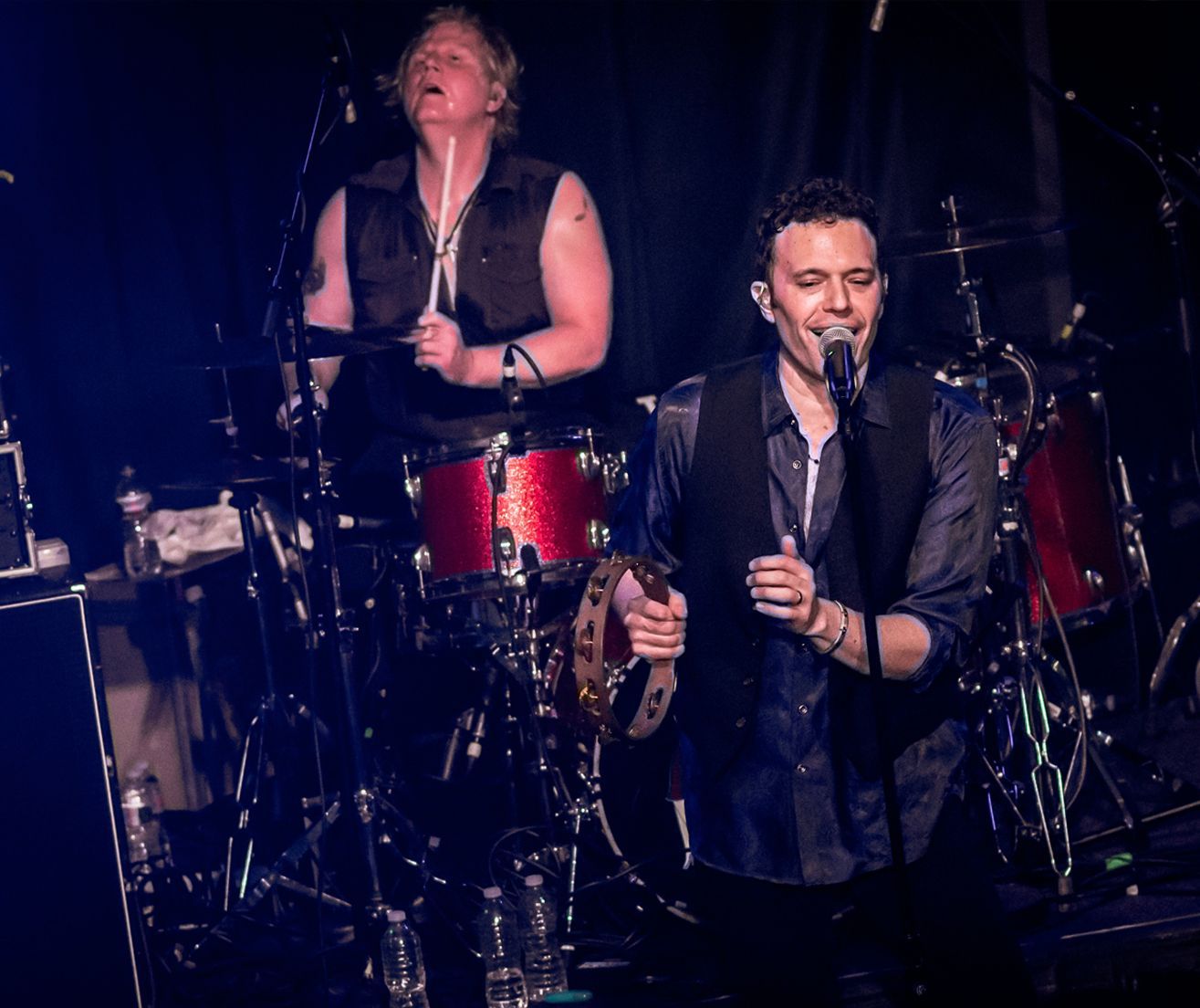 Two musicians performing on stage: a drummer and a singer with a tambourine.