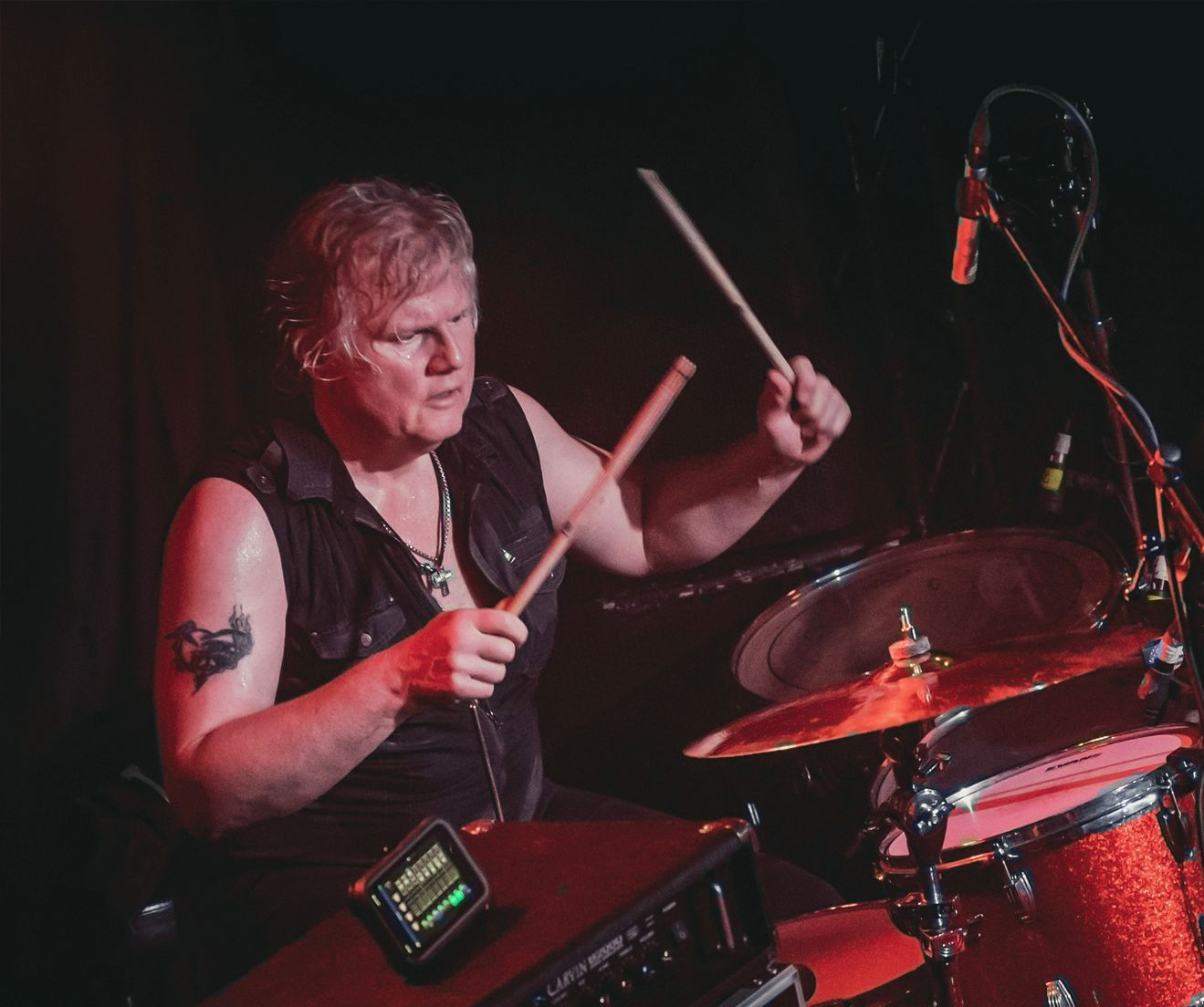 Drummer playing on stage, hitting cymbals. Red-lit stage, dark clothing, tattoo on arm.