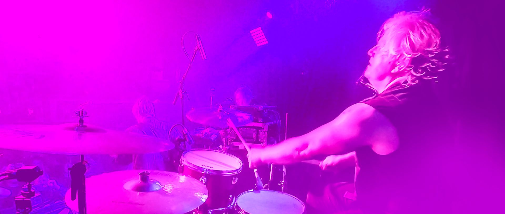 Drummer performing on stage, bathed in purple and pink stage lighting.
