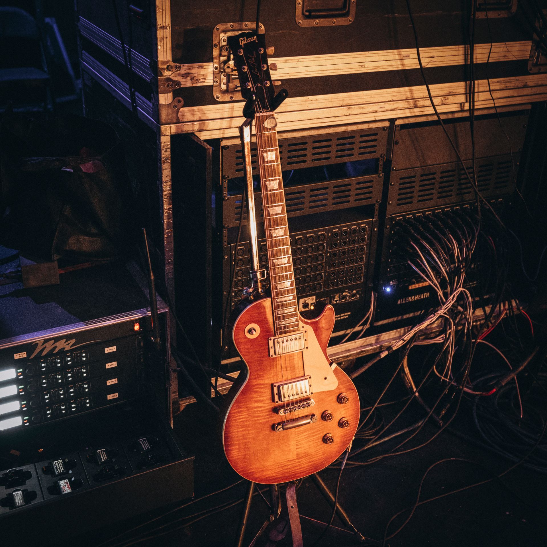 A brown electric guitar on a stand backstage. Cables and equipment surround it.