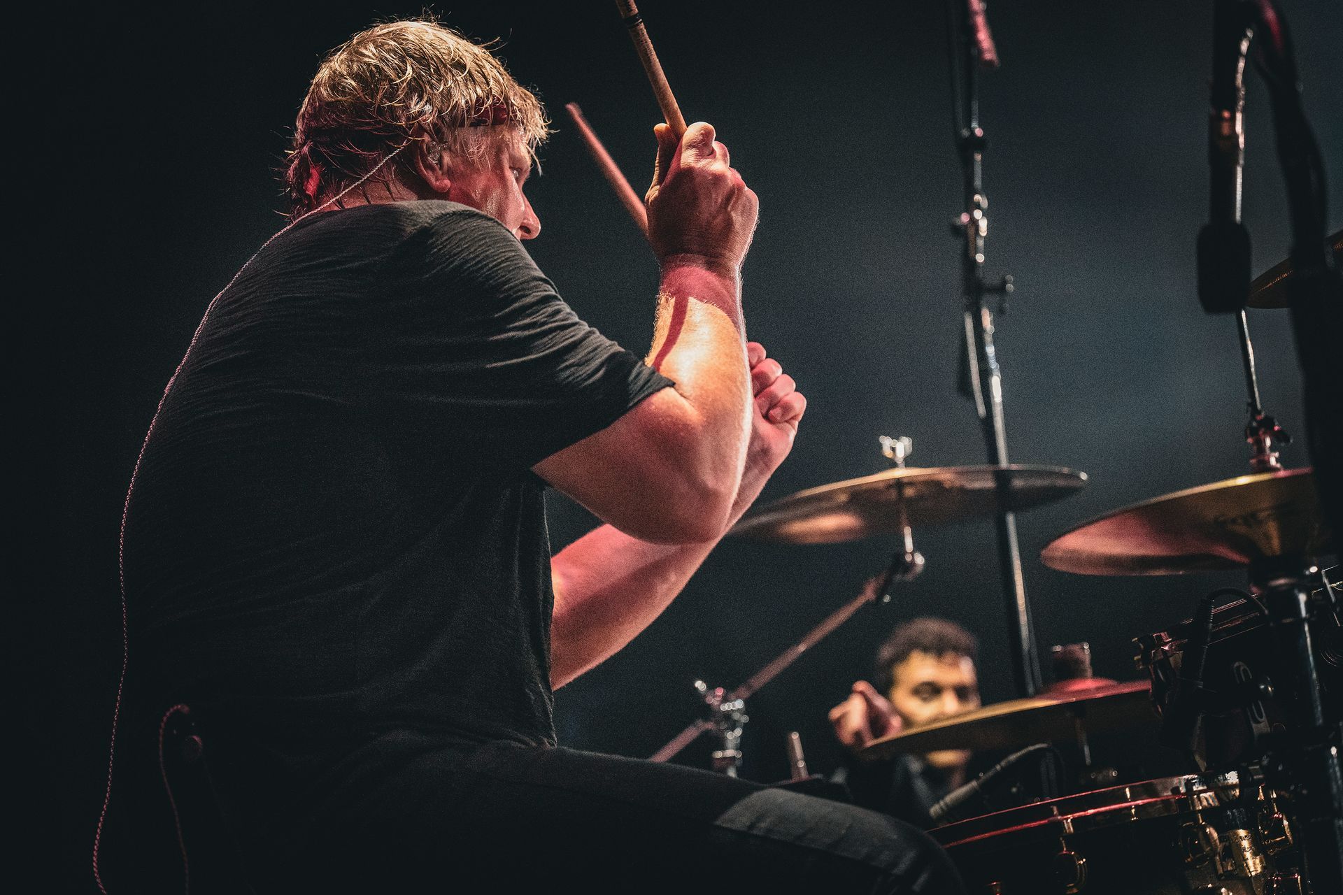 Drummer playing a drum kit on stage with another drummer visible in the background.