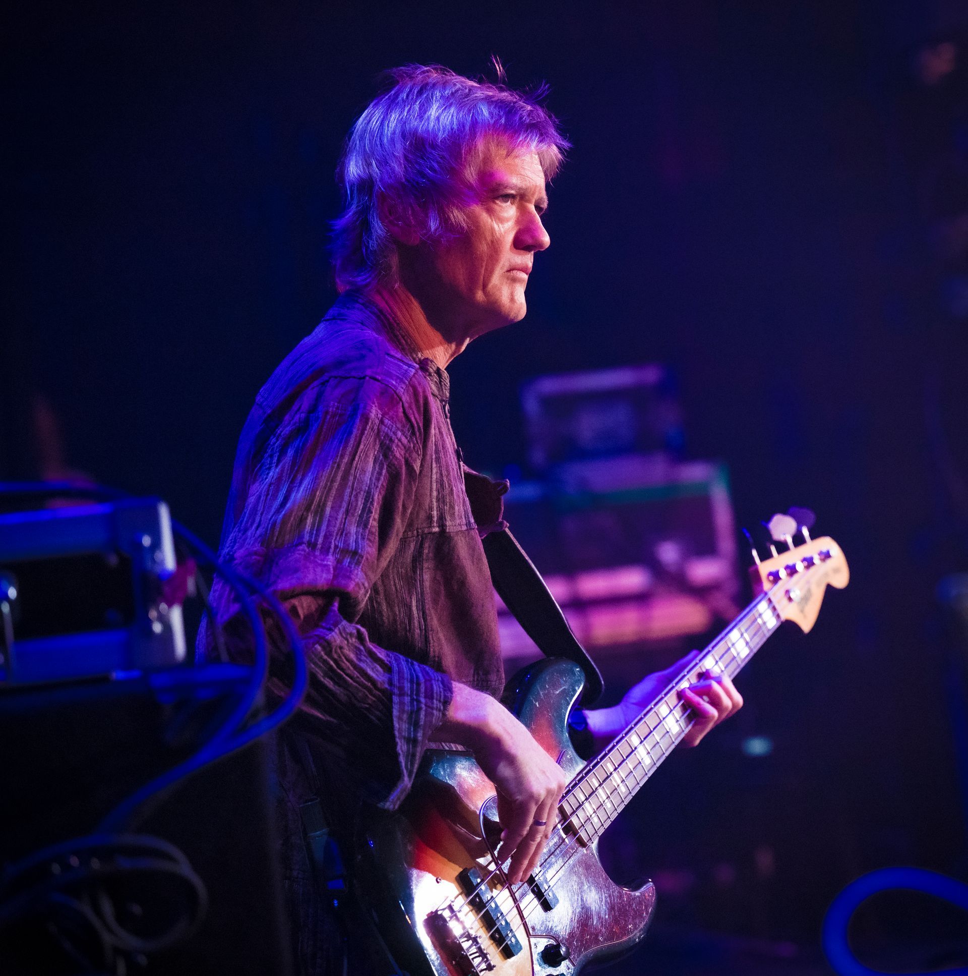 A musician playing a bass guitar on stage with blue and purple lighting.