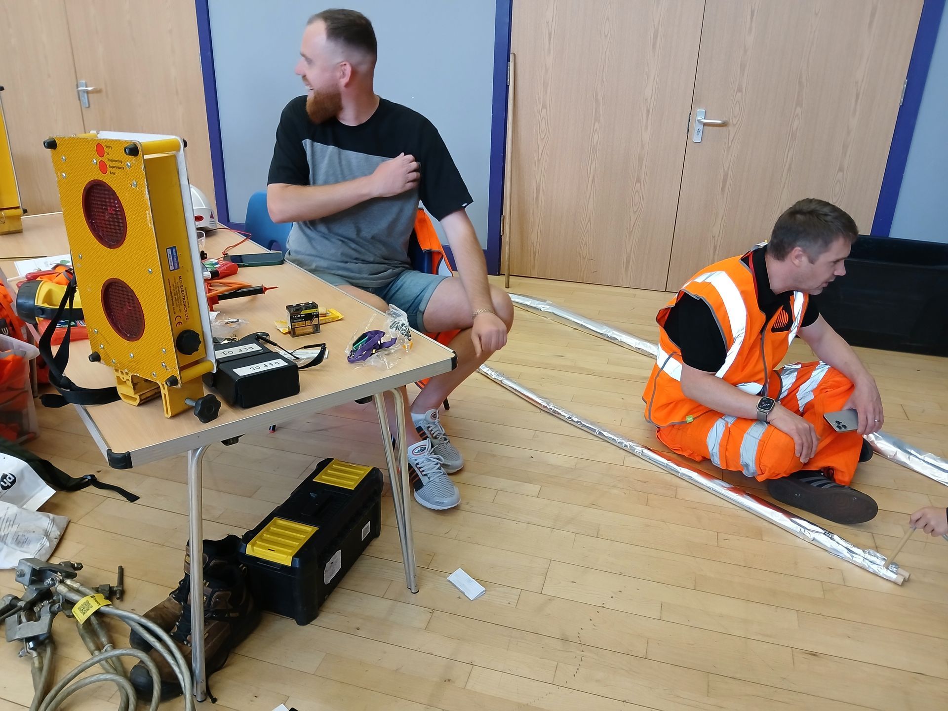Two men in a room; one at a table with tools and equipment, the other seated near metal bars, both wearing work clothing.