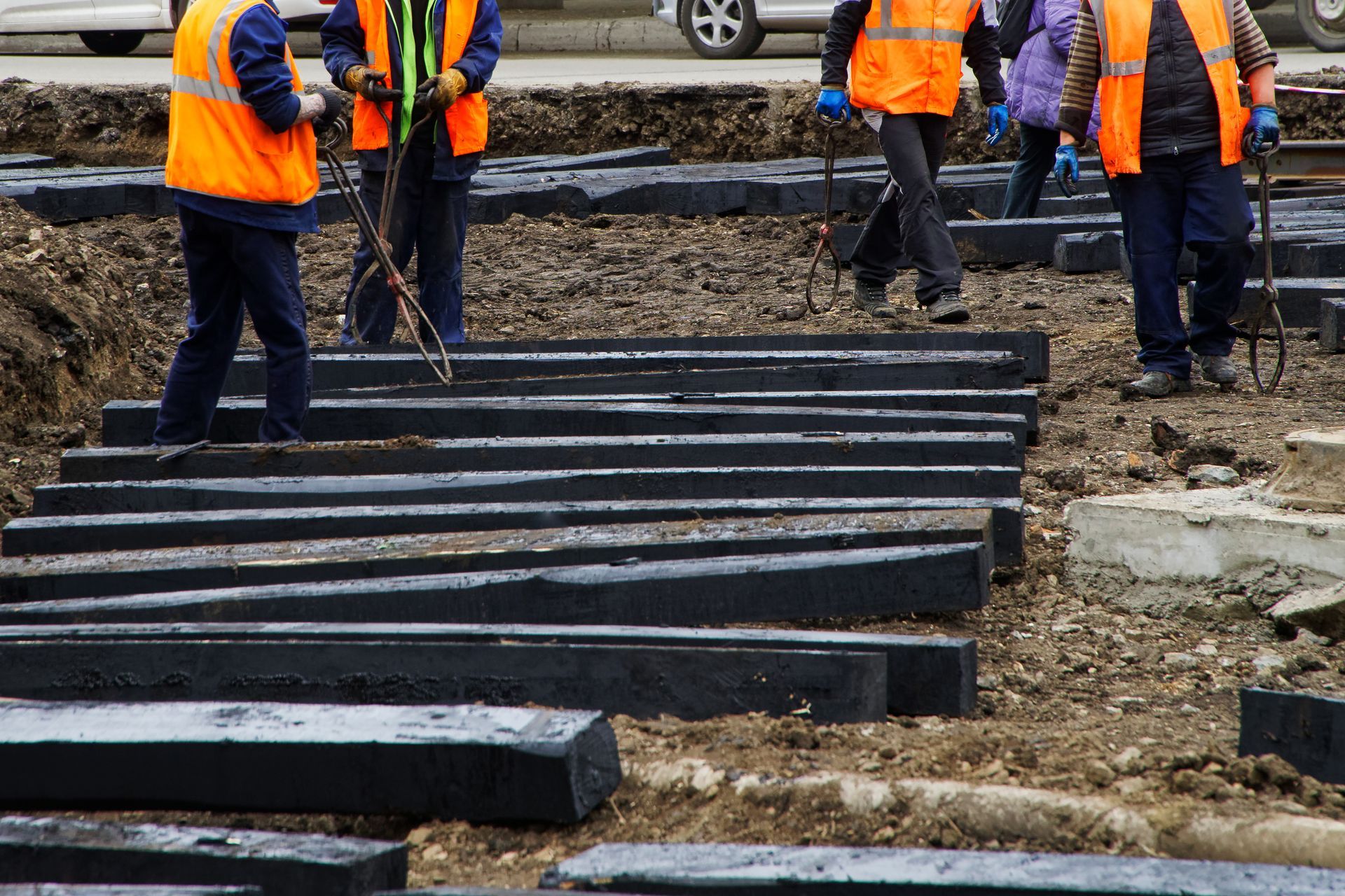 Workers in orange vests install dark, rectangular paving stones. Dirt and construction site visible.