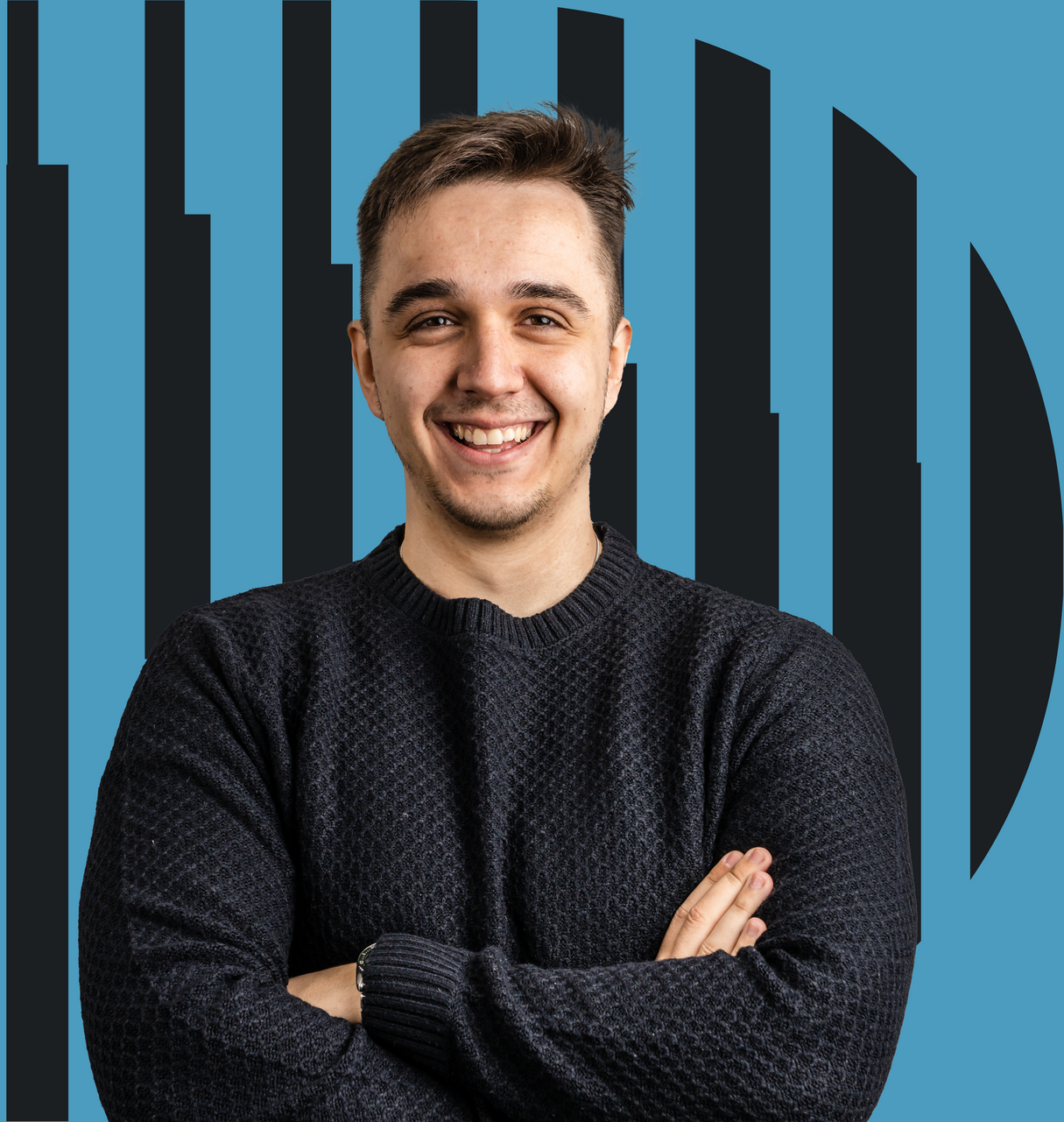 Smiling man with arms crossed, in a dark sweater, in front of a blue background with black vertical stripes.