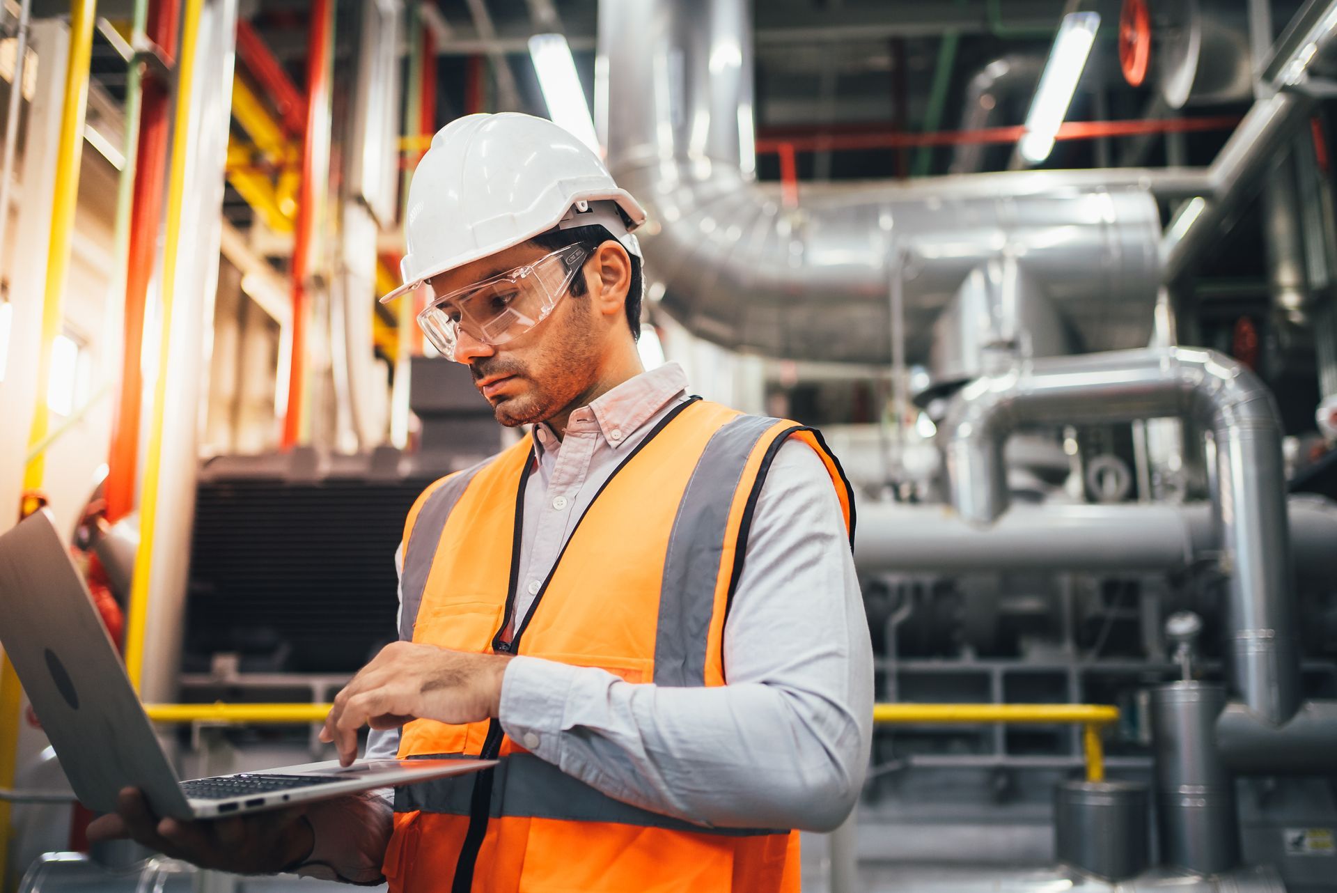 Engineer in safety gear uses a laptop in an industrial setting, focused expression.