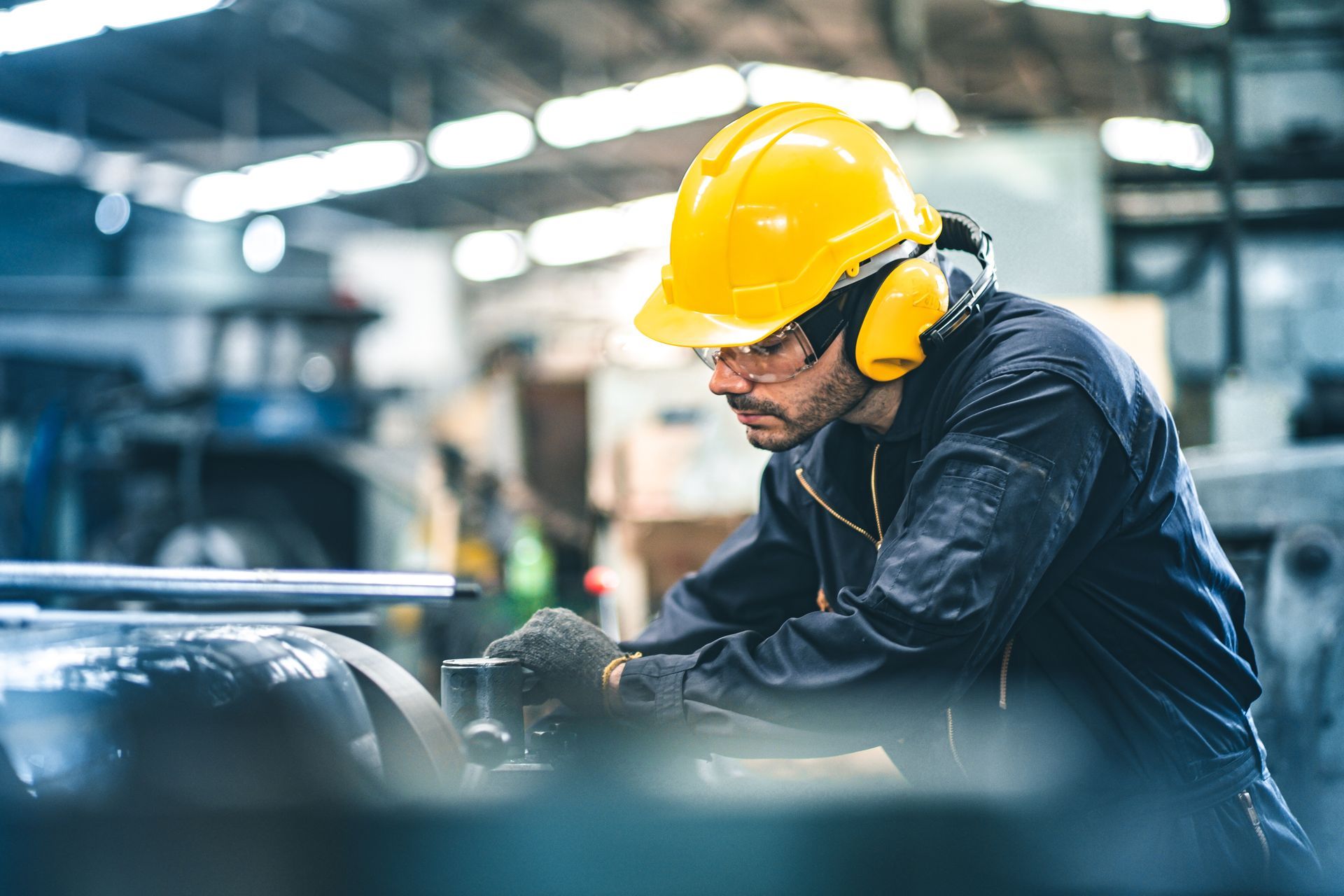 Worker in a yellow hard hat and hearing protection operates machinery in a factory.