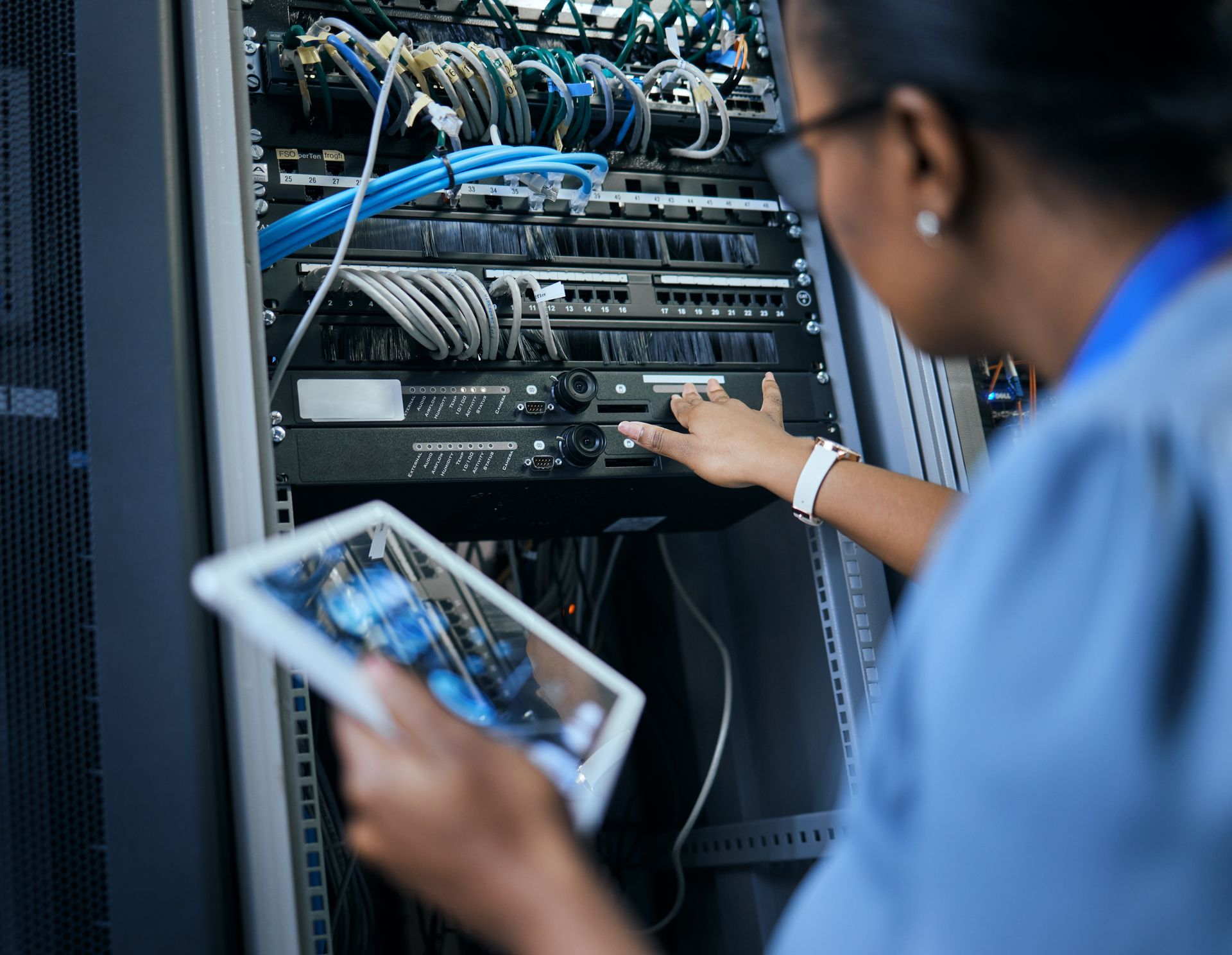 Technician in blue shirt uses a tablet while working on a server rack filled with cables and devices.