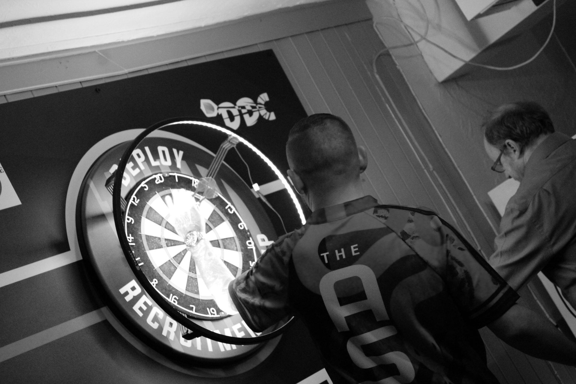 A person throws a dart at a dartboard, another watches. Game in progress, indoor setting.