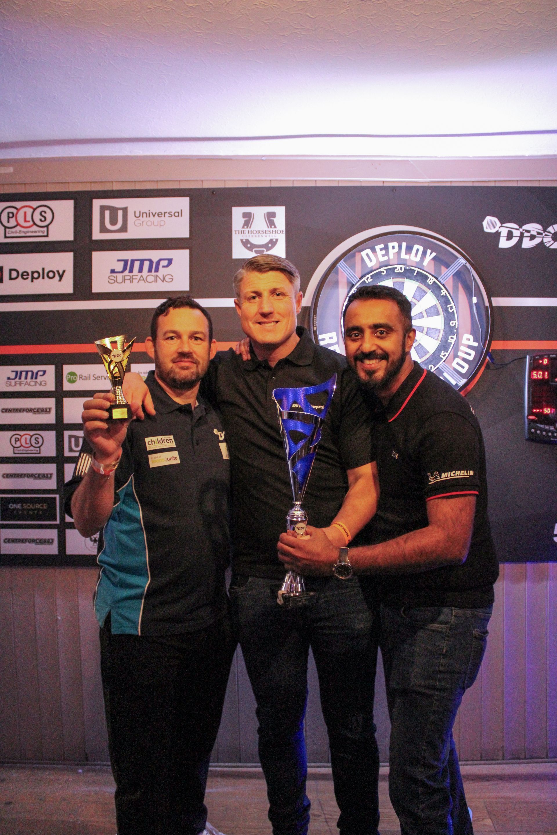 Three men celebrate a darts victory, holding trophies in front of a dartboard.