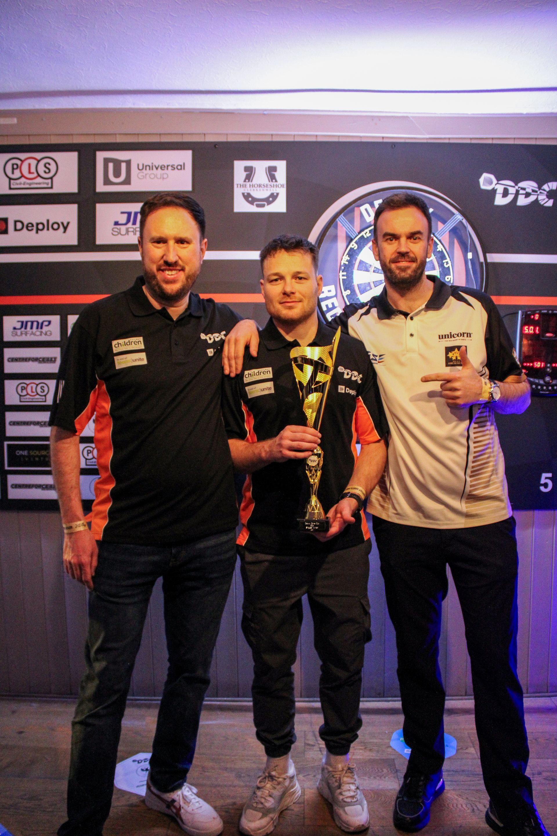 Three men pose with a trophy in front of a darts board. One holds the trophy.