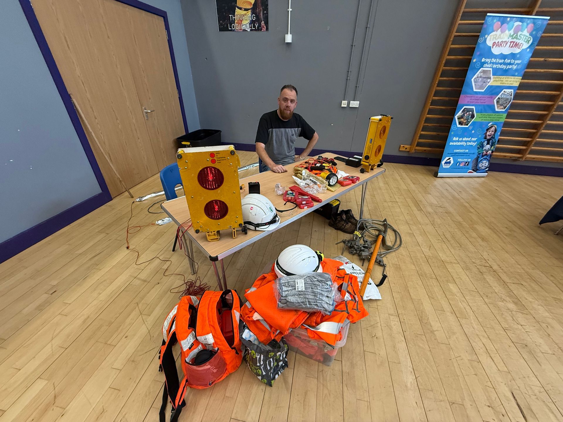 Man at table with safety gear. Yellow boxes, orange vests, hard hats, ropes, setting: indoor, wood floor.