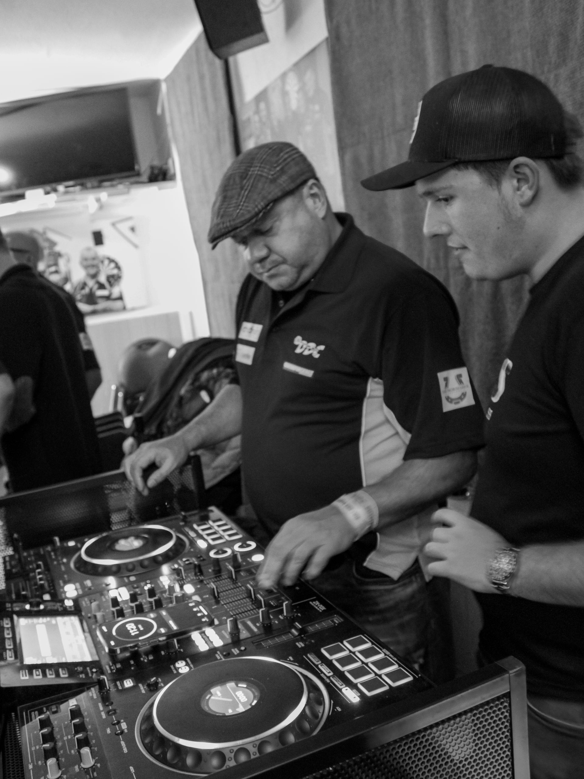 Two men operating DJ equipment indoors, one wearing a flat cap.