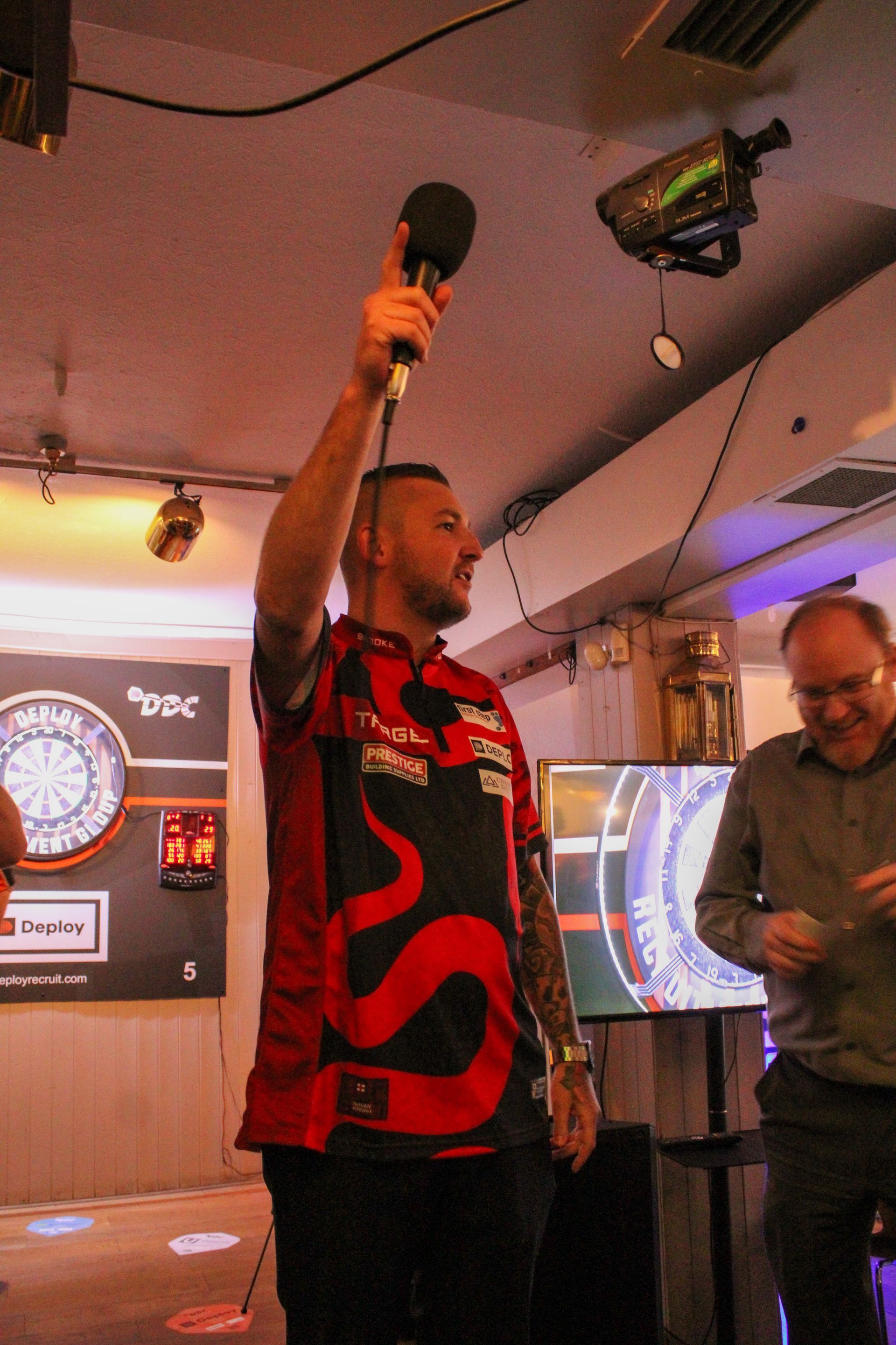 Man holding microphone, arm raised. Wearing red shirt with black accents, darts setting. Another man watches.