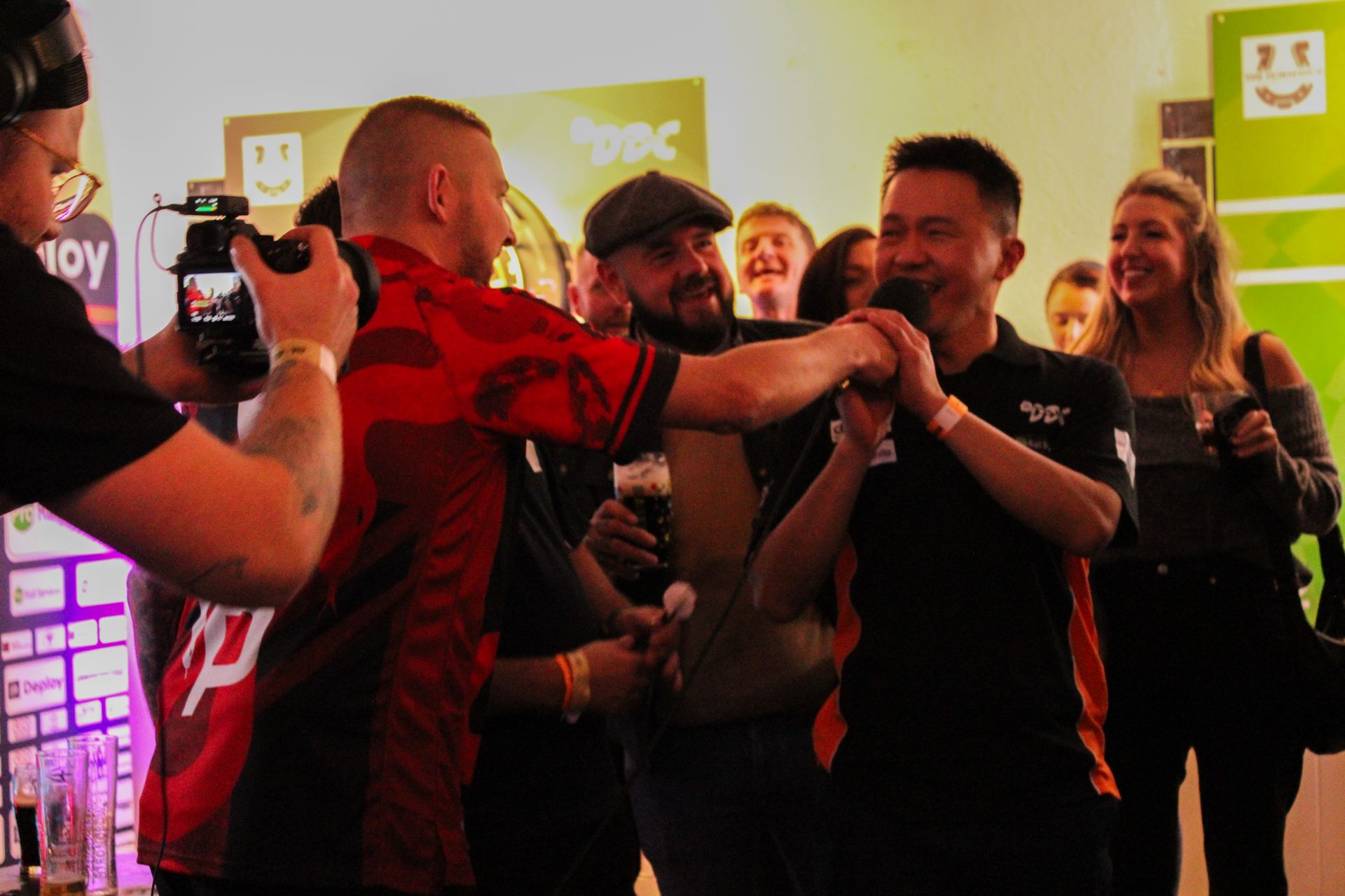Man in red jersey high-fives another holding a microphone. Other people watch in a pub-like setting.