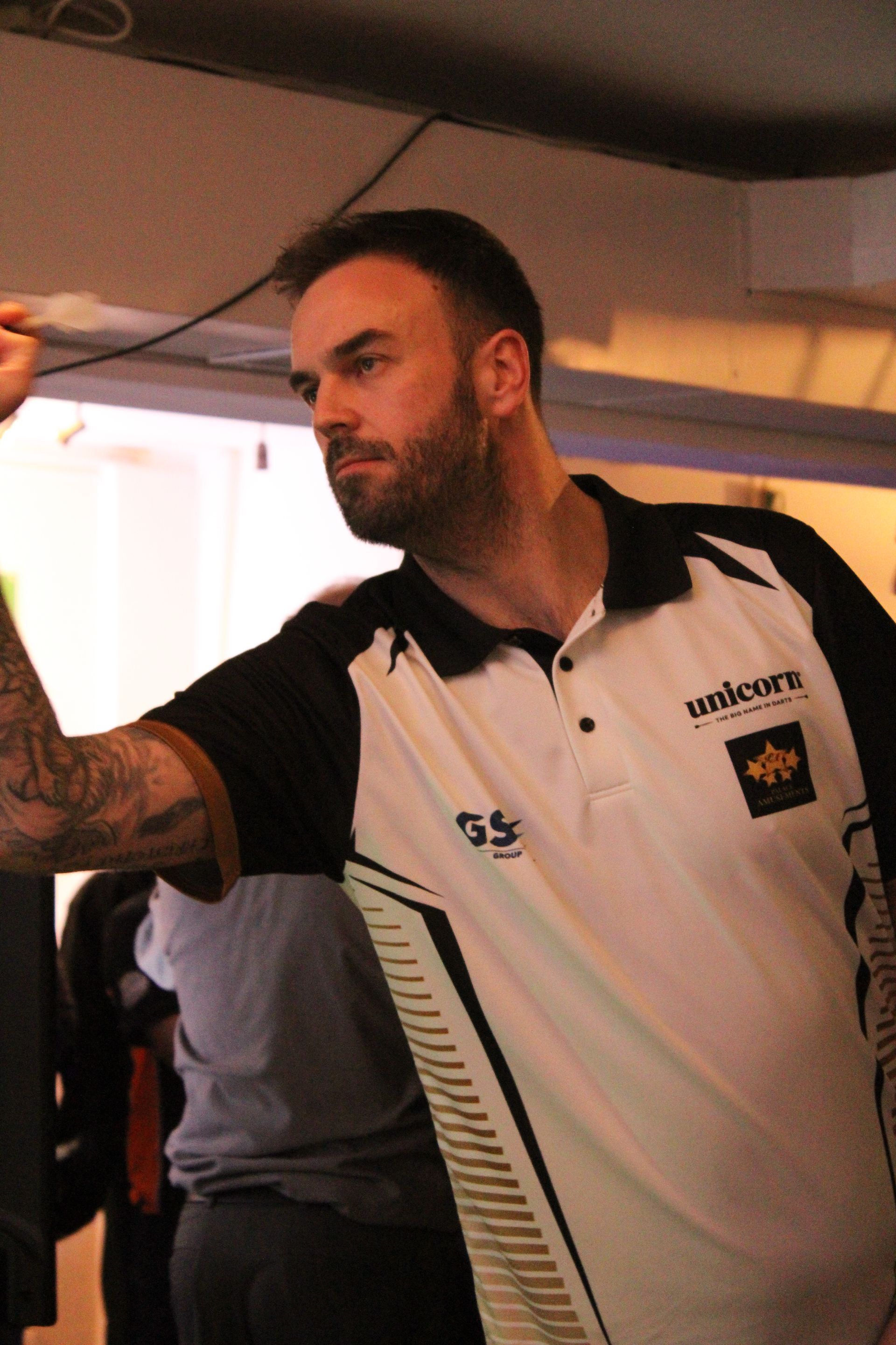 Man with a beard throwing a dart, wearing a white and black collared shirt, inside.