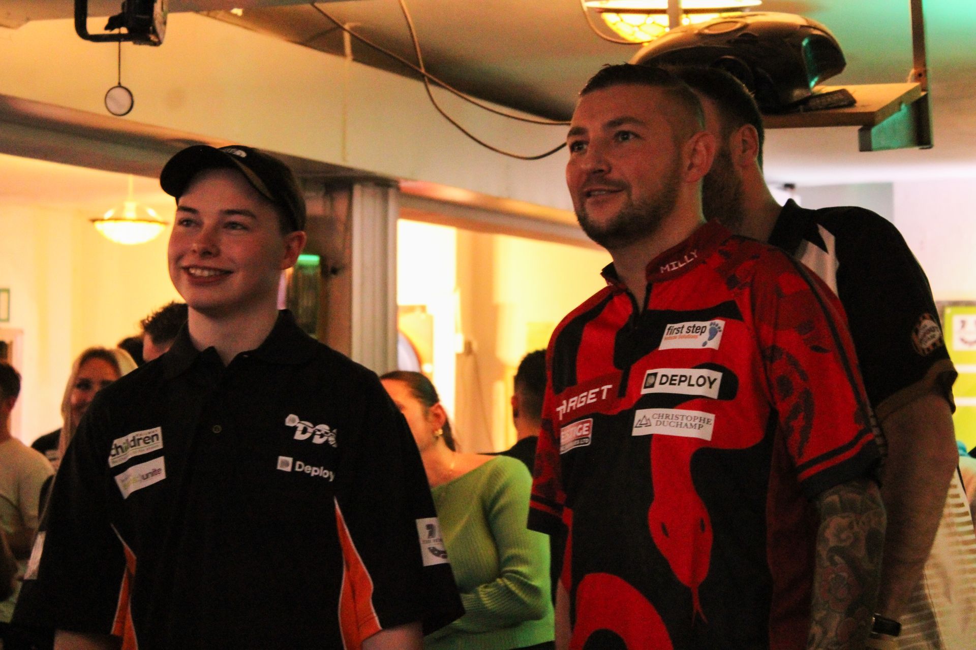 Two dart players smiling at a dart match. One in black, one in red, in a bar setting.