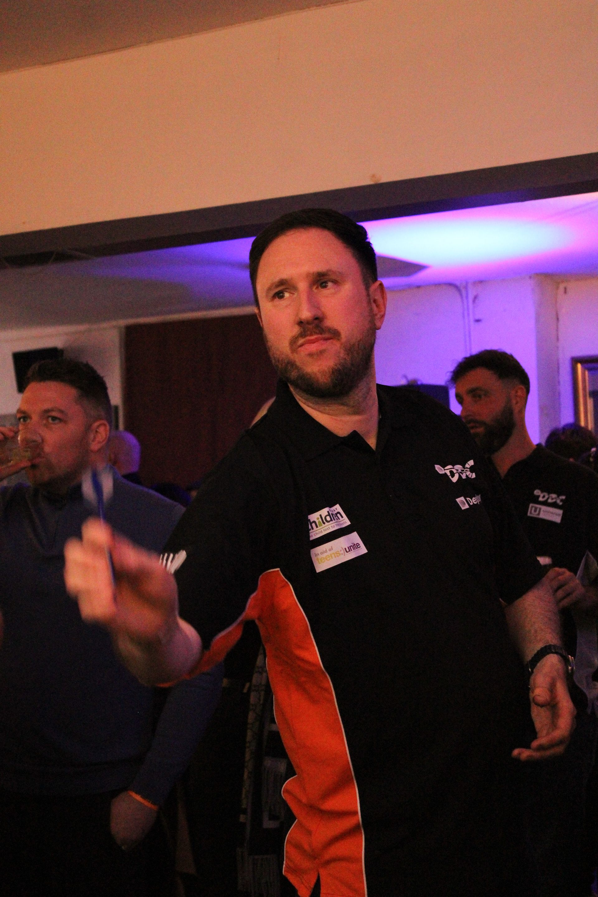 Man in black shirt throws a dart, other men in pub setting. Orange and purple lighting.