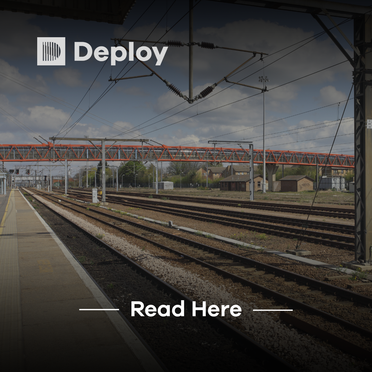 Train tracks at a railway station with overhead wires and a pedestrian bridge.