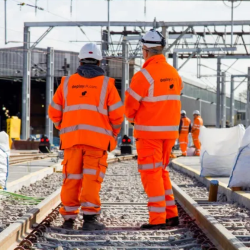 Two workers in orange safety gear stand on railway tracks, observing construction.