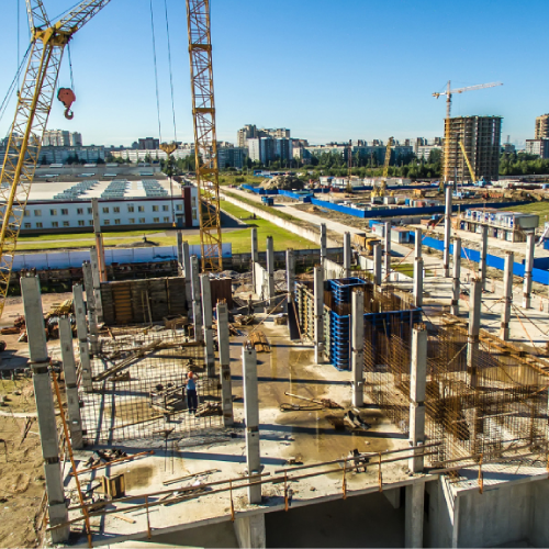 Construction site with cranes, concrete pillars, and partially built structure; city in the background.