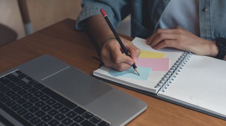 Hands writing in a spiral notebook beside a laptop on a wooden desk with colorful sticky notes.