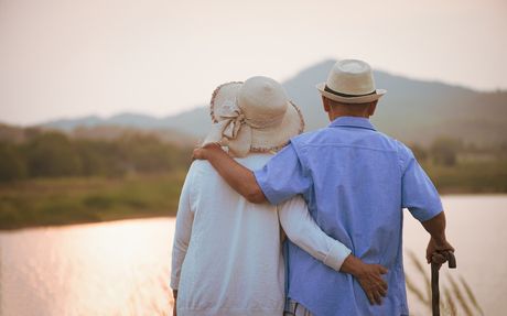 Elderly couple walking arm in arm by a lakeside at sunset, with hats and a relaxed, warm scene