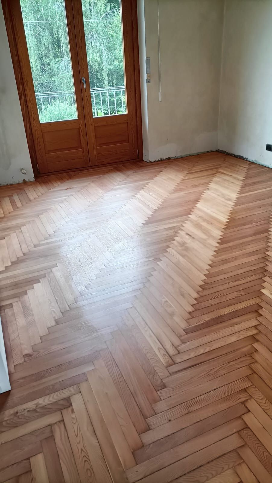 Stanza vuota con pavimento in legno a spina di pesce e porte in legno e vetro che conducono all'esterno.