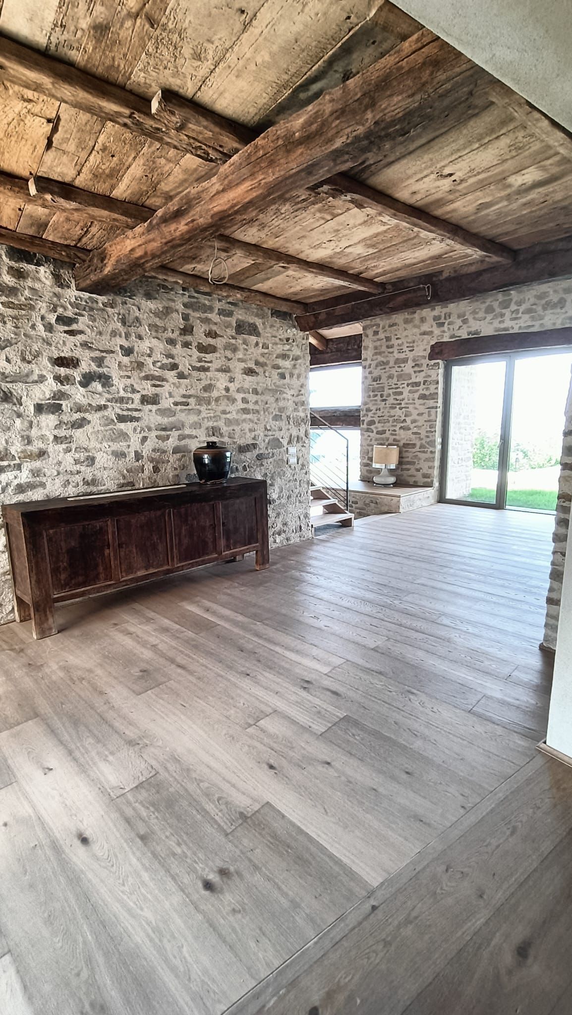Stanza rustica in pietra con travi in ​​legno a vista, pavimento in assi di legno bianco e un armadio in legno scuro accanto alle grandi finestre.