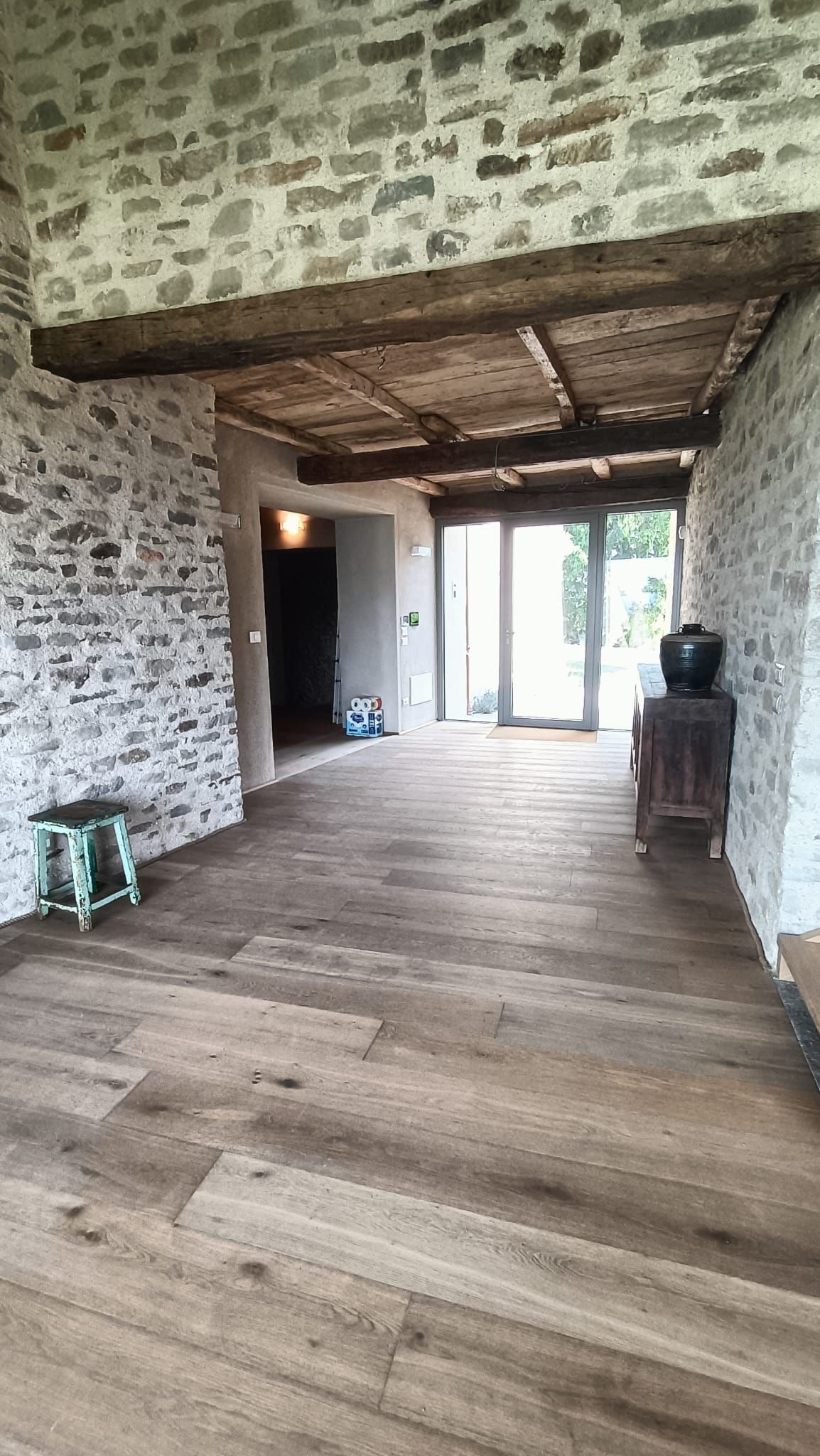 Corridoio con pareti in pietra, pavimento in assi di legno e soffitto con travi a vista, che conduce a una luminosa porta.