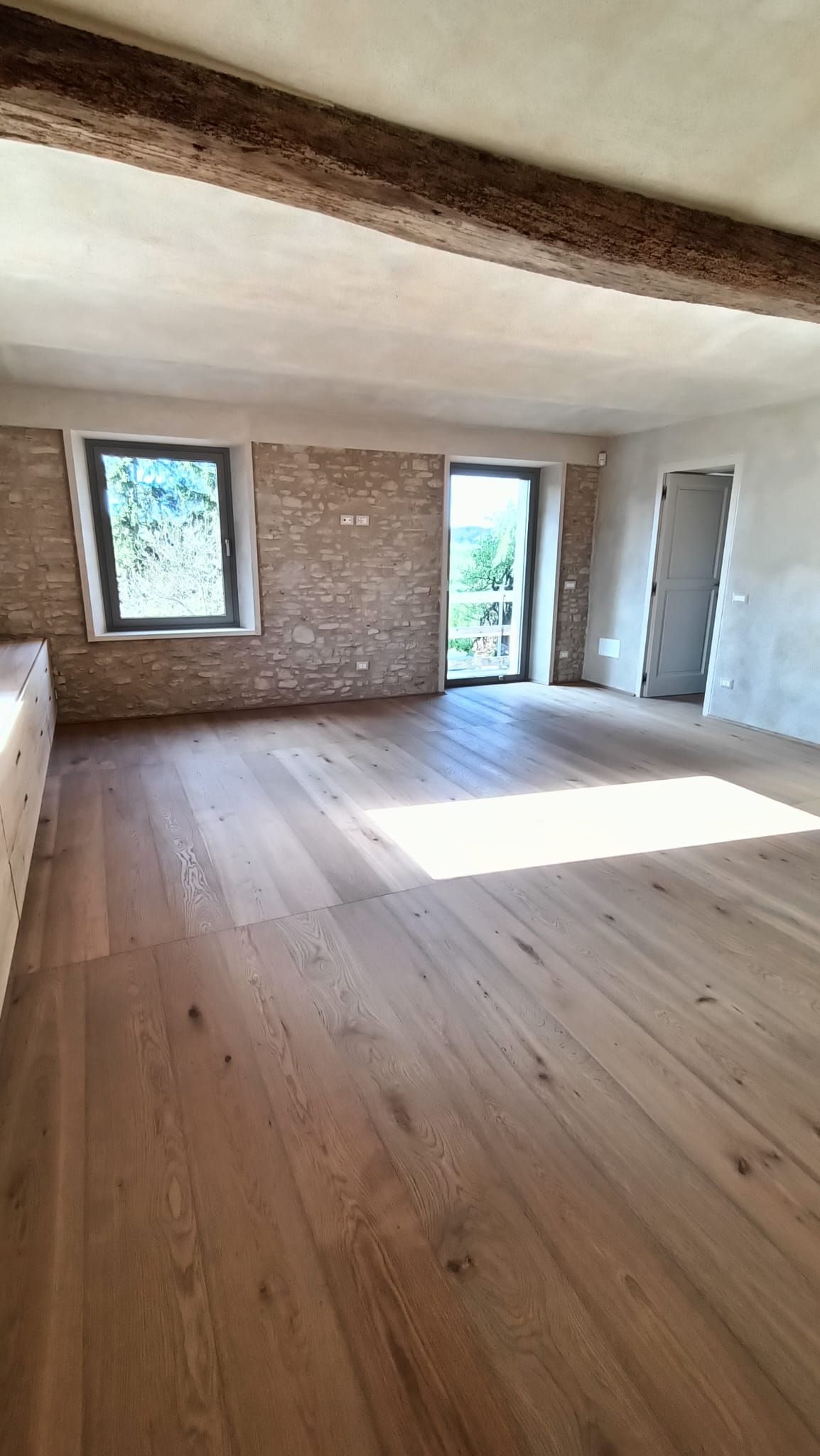 Stanza rustica vuota con pavimento in legno, travi a vista, due finestre e una zona soleggiata sul pavimento.