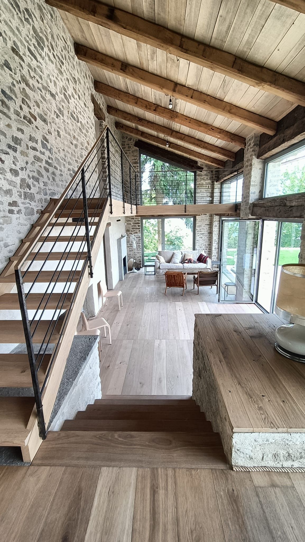 Ampio loft rustico con travi a vista, scala e grandi finestre che si affacciano su una terrazza.