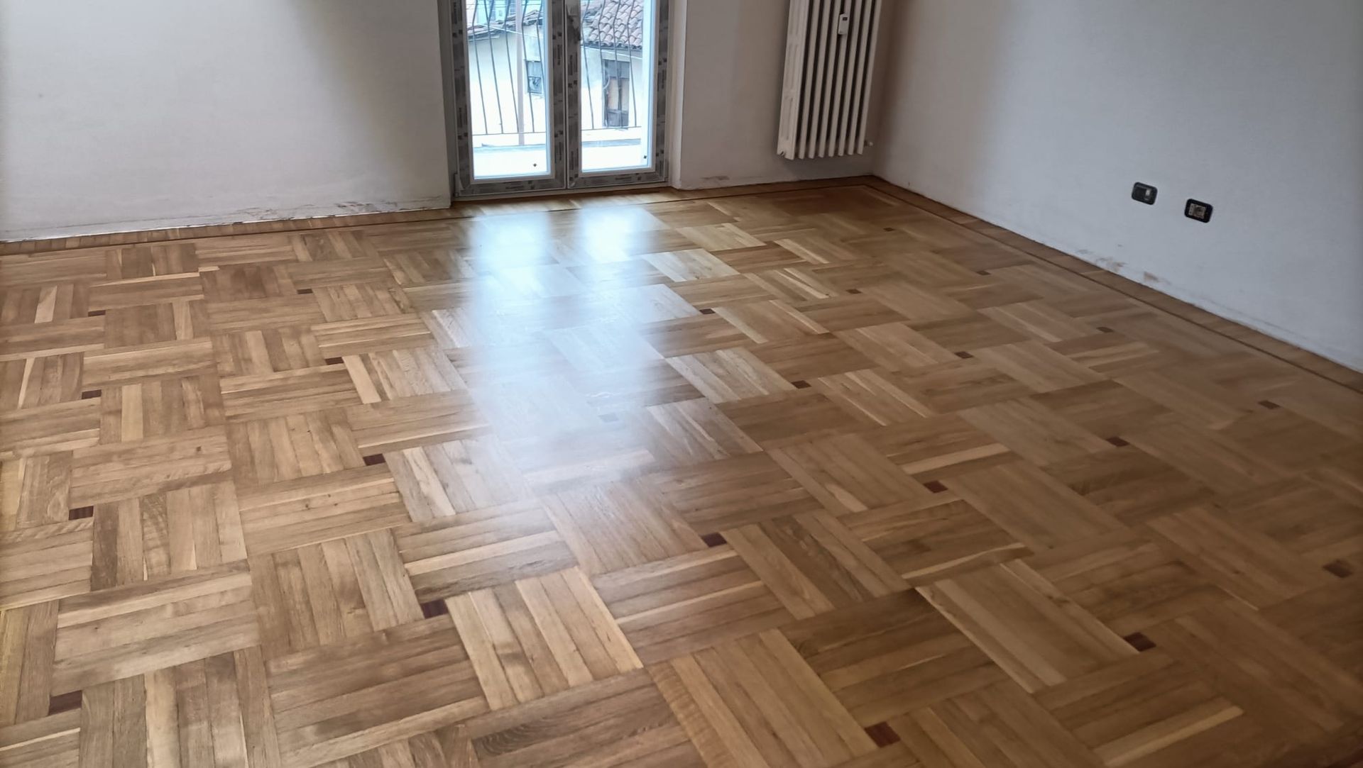 Stanza vuota con pavimento in legno a spina di pesce, pareti bianche e porte finestre che si aprono su un balcone.