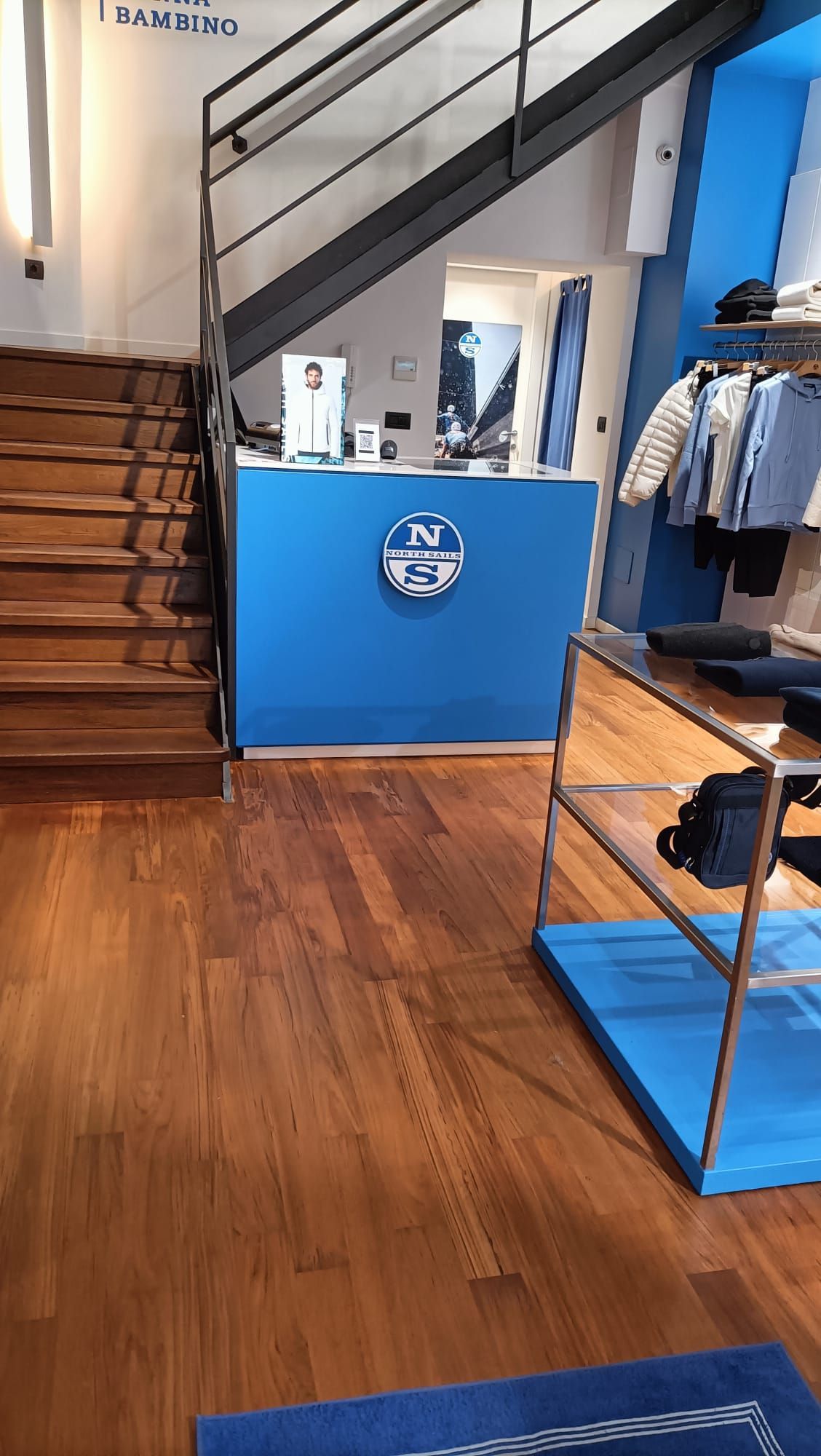 Interno di un negozio di abbigliamento con pavimento in legno, scala, bancone blu e appendiabiti.