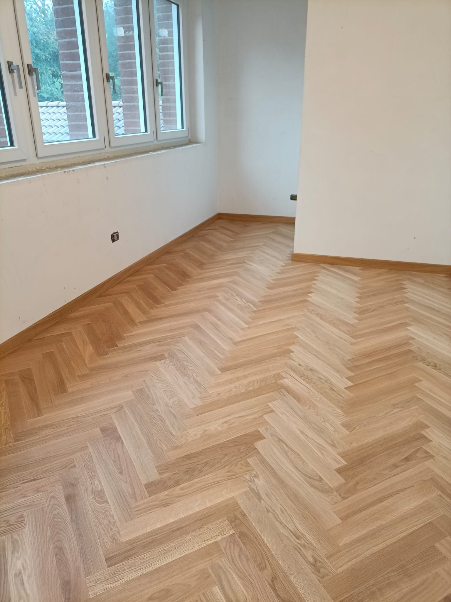 Stanza vuota con pavimento in legno a spina di pesce, pareti bianche e finestre sulla sinistra.