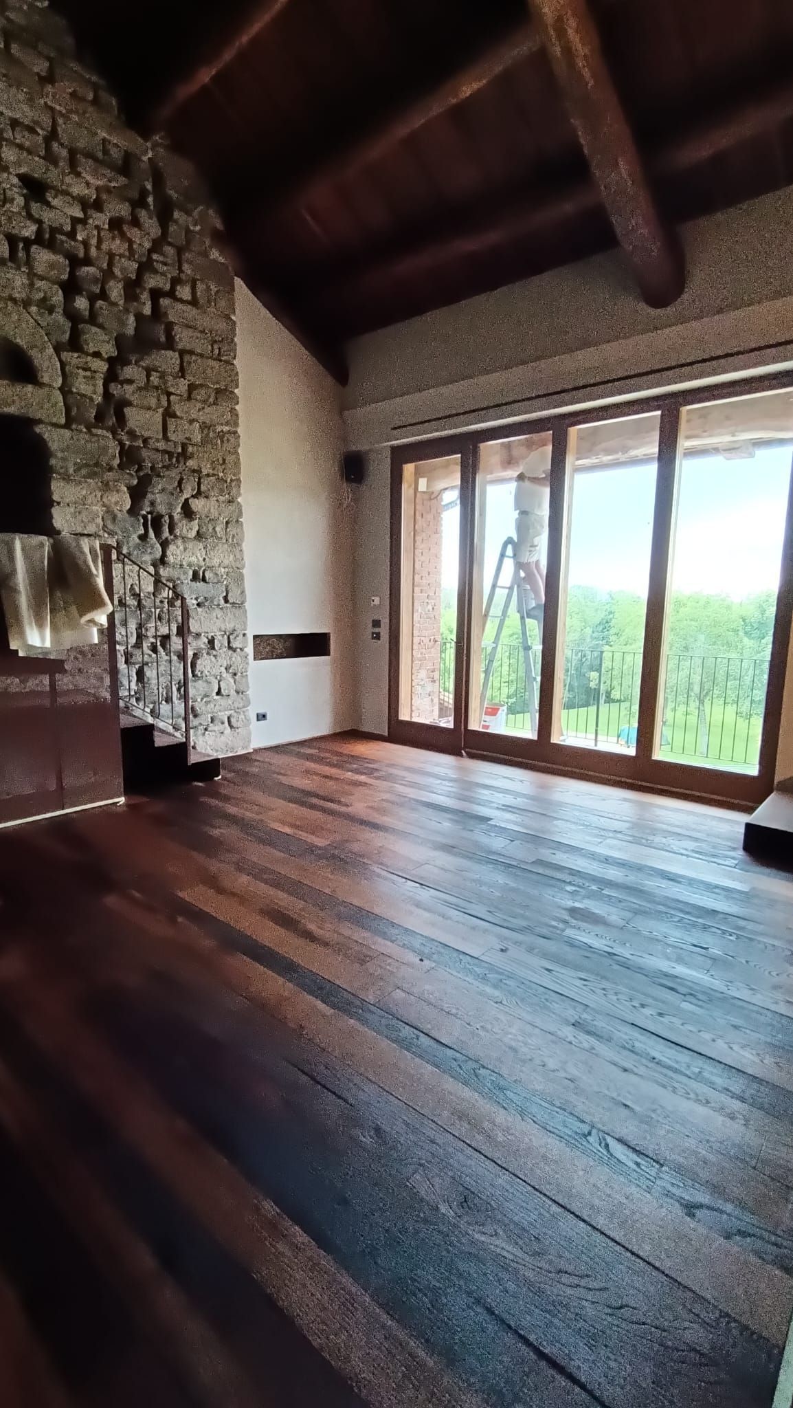 Camera rustica con pavimento in legno scuro, parete in pietra, travi a vista e grandi porte finestre che si affacciano sul verde.