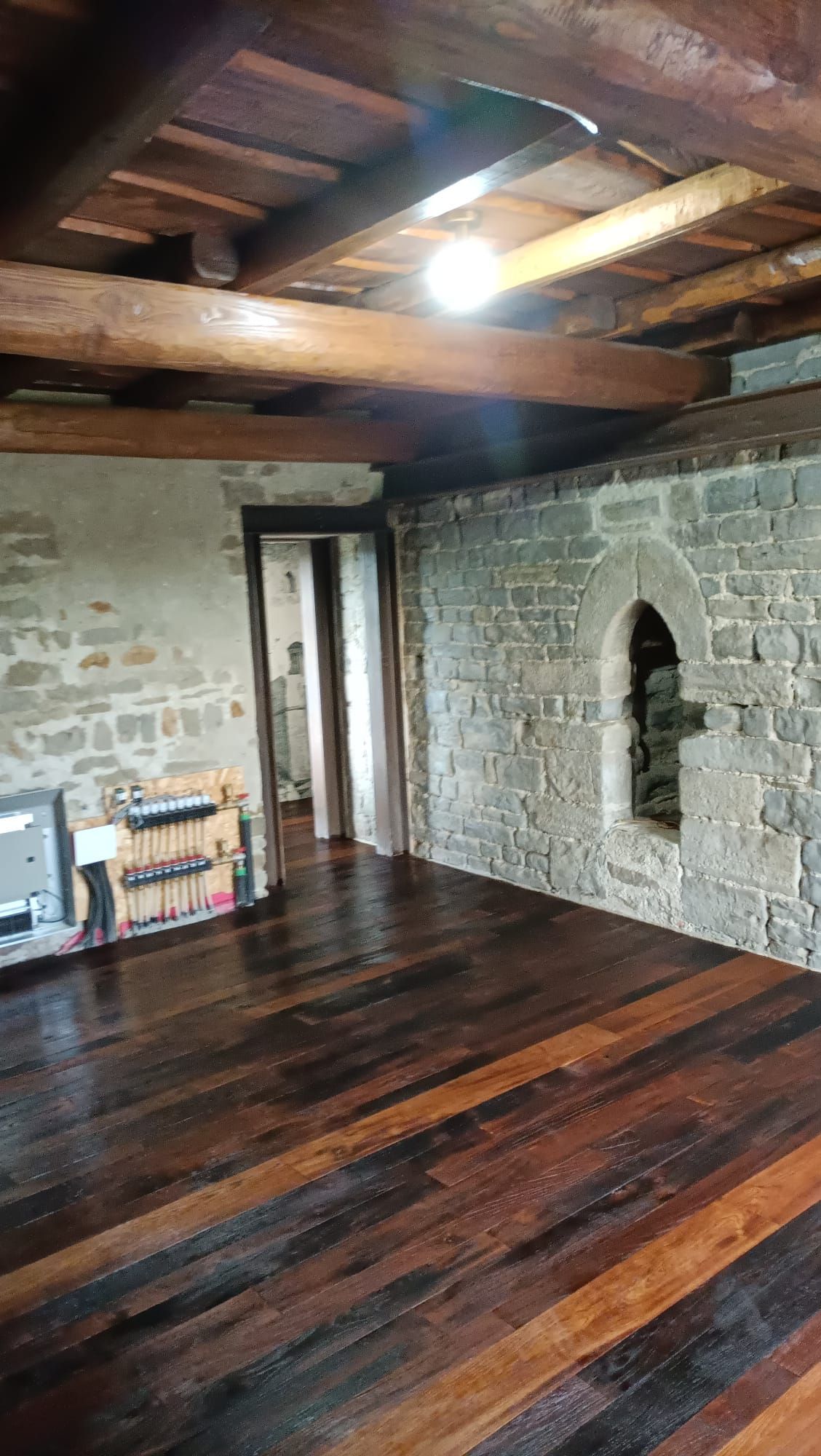 Camera rustica con pavimento e soffitto in legno scuro, parete del camino in pietra e una porta luminosa.