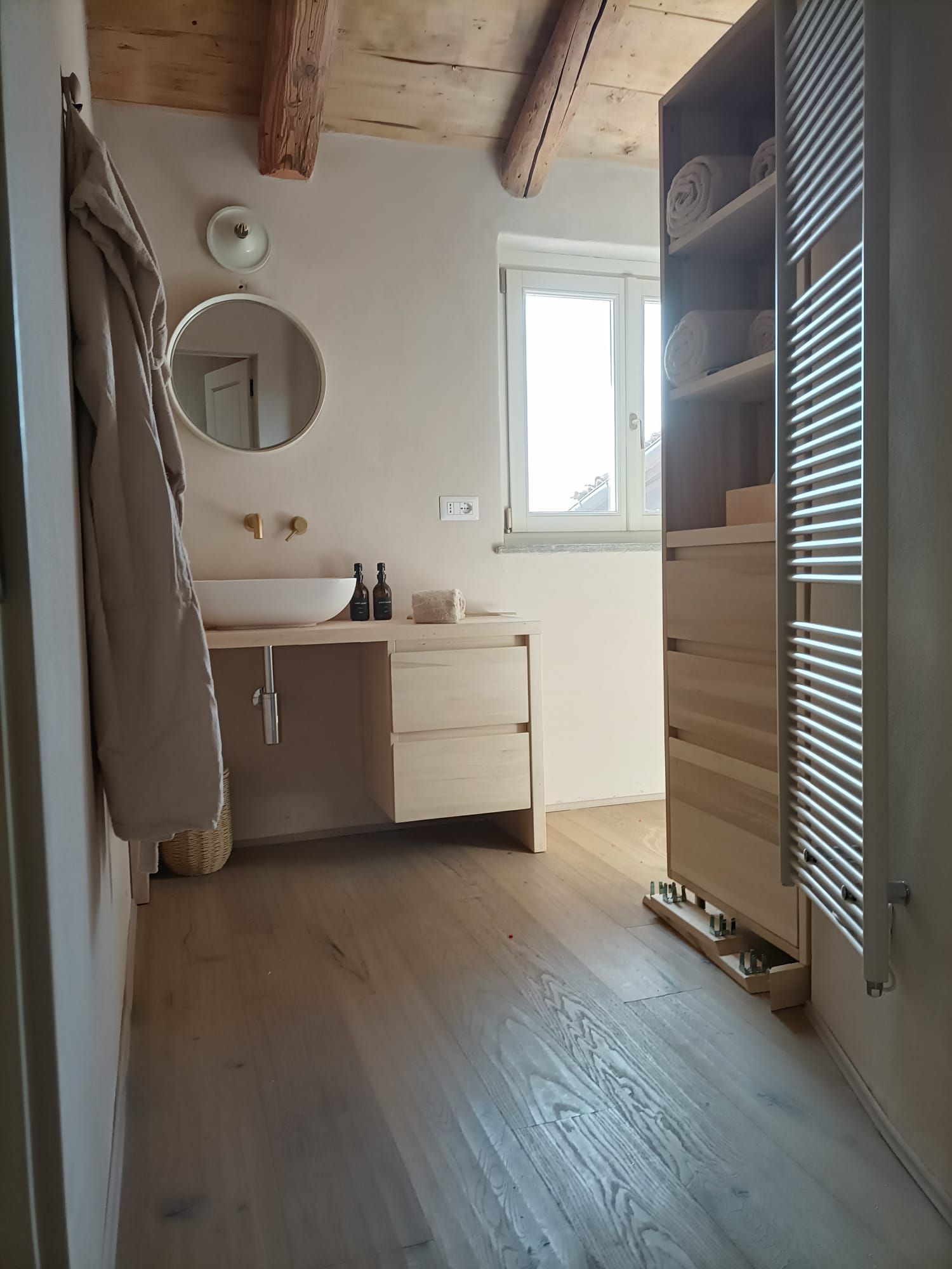 Bagno luminoso con mobile lavabo bianco, specchio rotondo, lucernario, travi in ​​legno e porta dell'armadio a doghe.